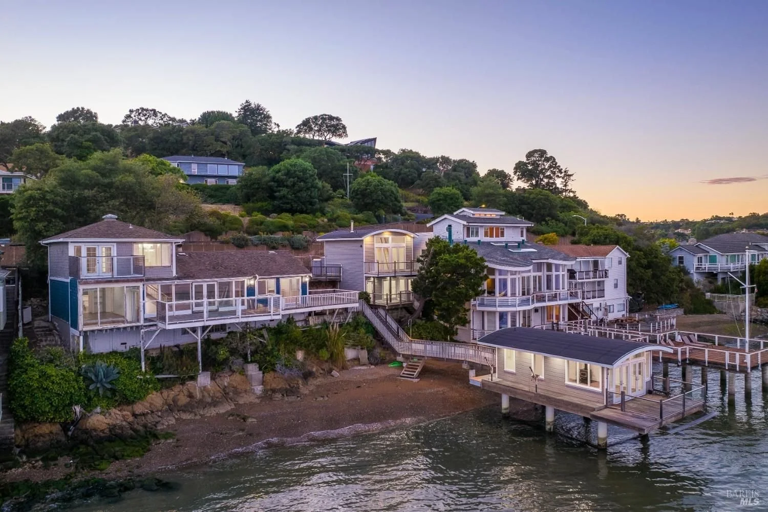 Waterfront houses with decks and staircases on a rocky shoreline at dusk, with green trees and hillside in the background.