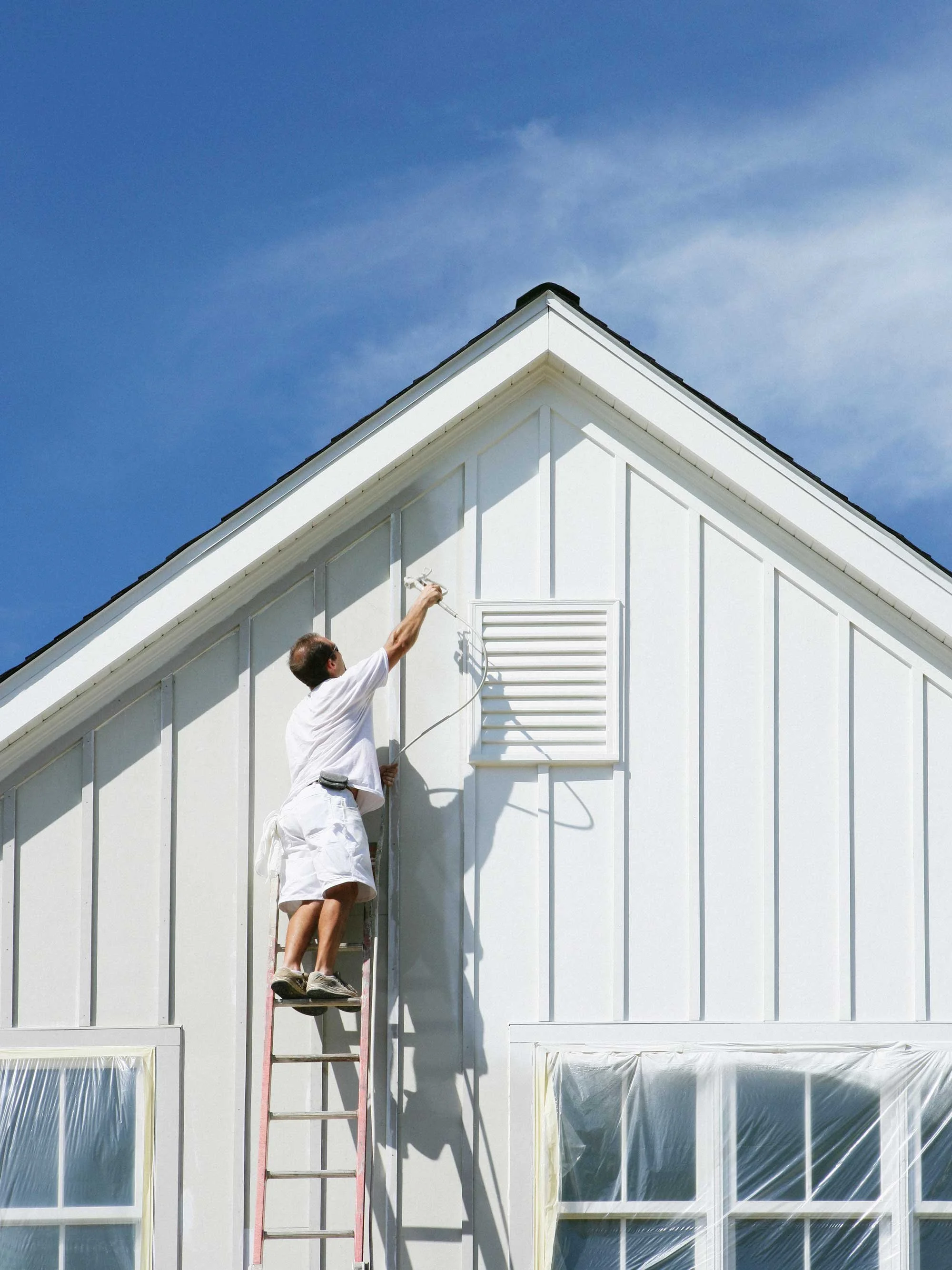 Man painting the exterior wall of a house while standing on a ladder, with a bright blue sky in the background.