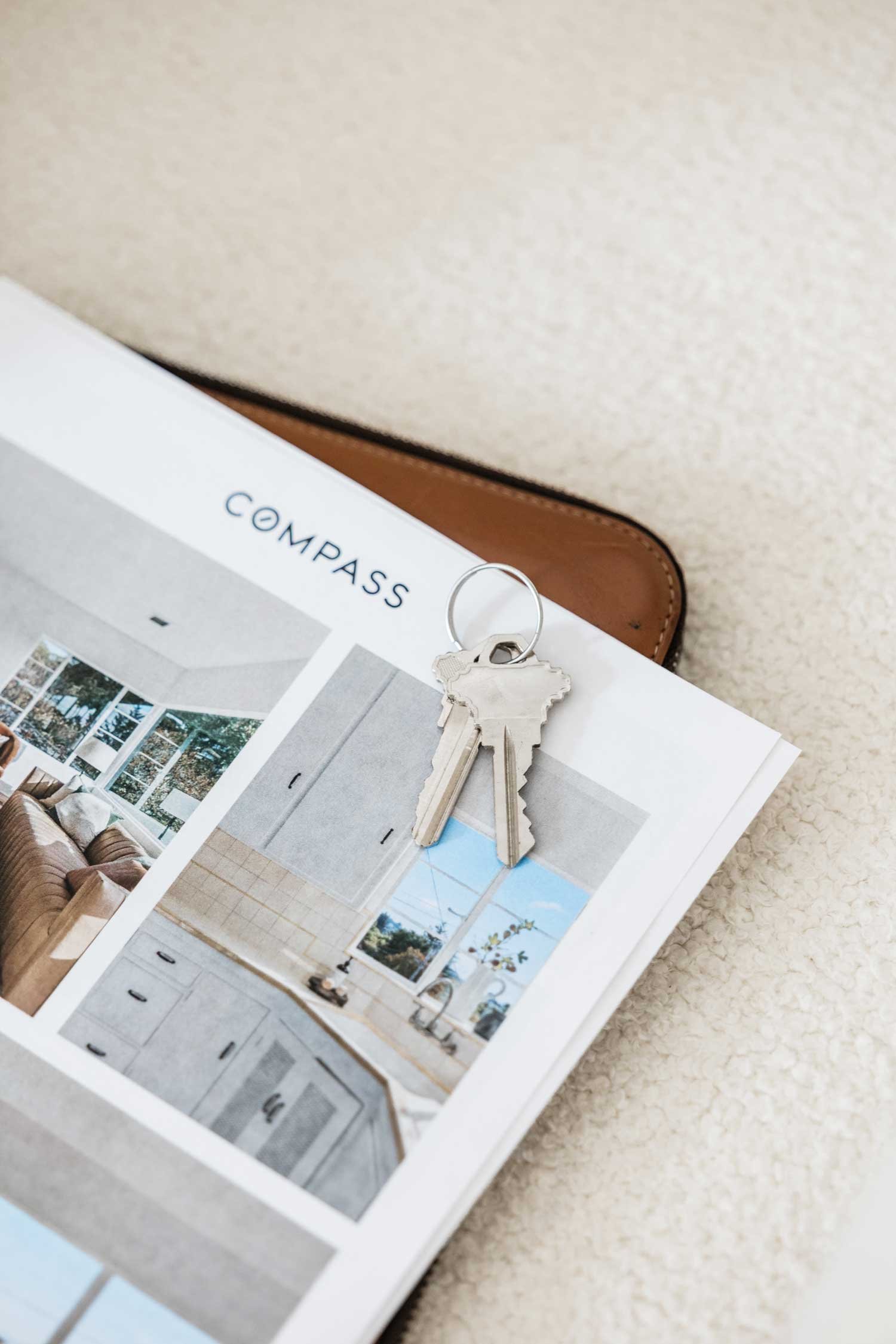 A set of keys resting on a printed interior design magazine titled 'COMPASS', with a brown leather case partially visible underneath, on a beige carpeted floor.