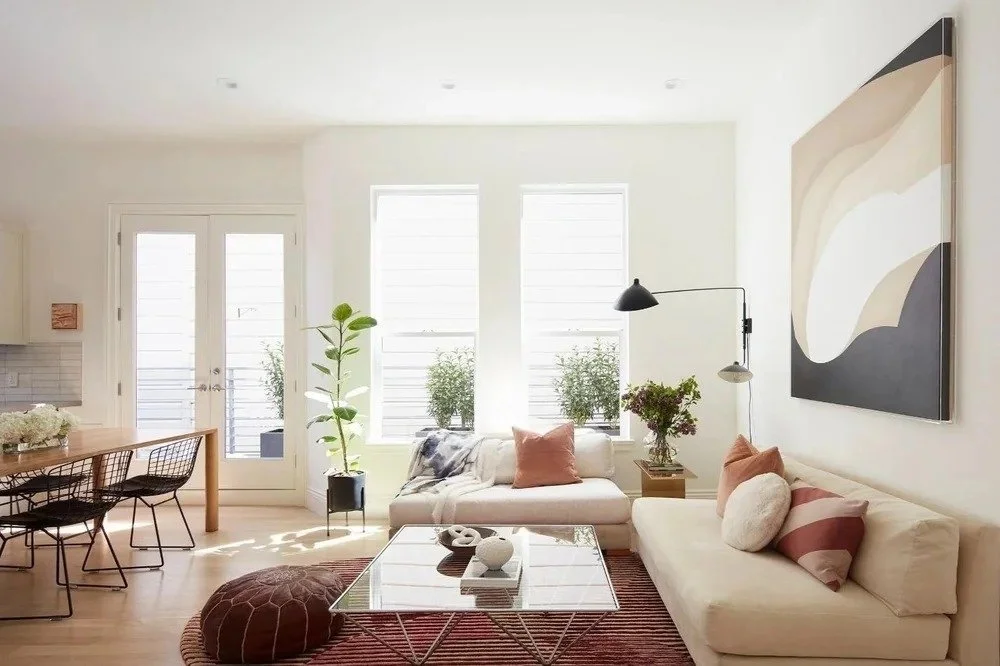 Bright living room with white walls, large windows, a white sofa with pillows, a glass coffee table, potted plants, a large abstract painting, and a wooden dining table with black chairs.