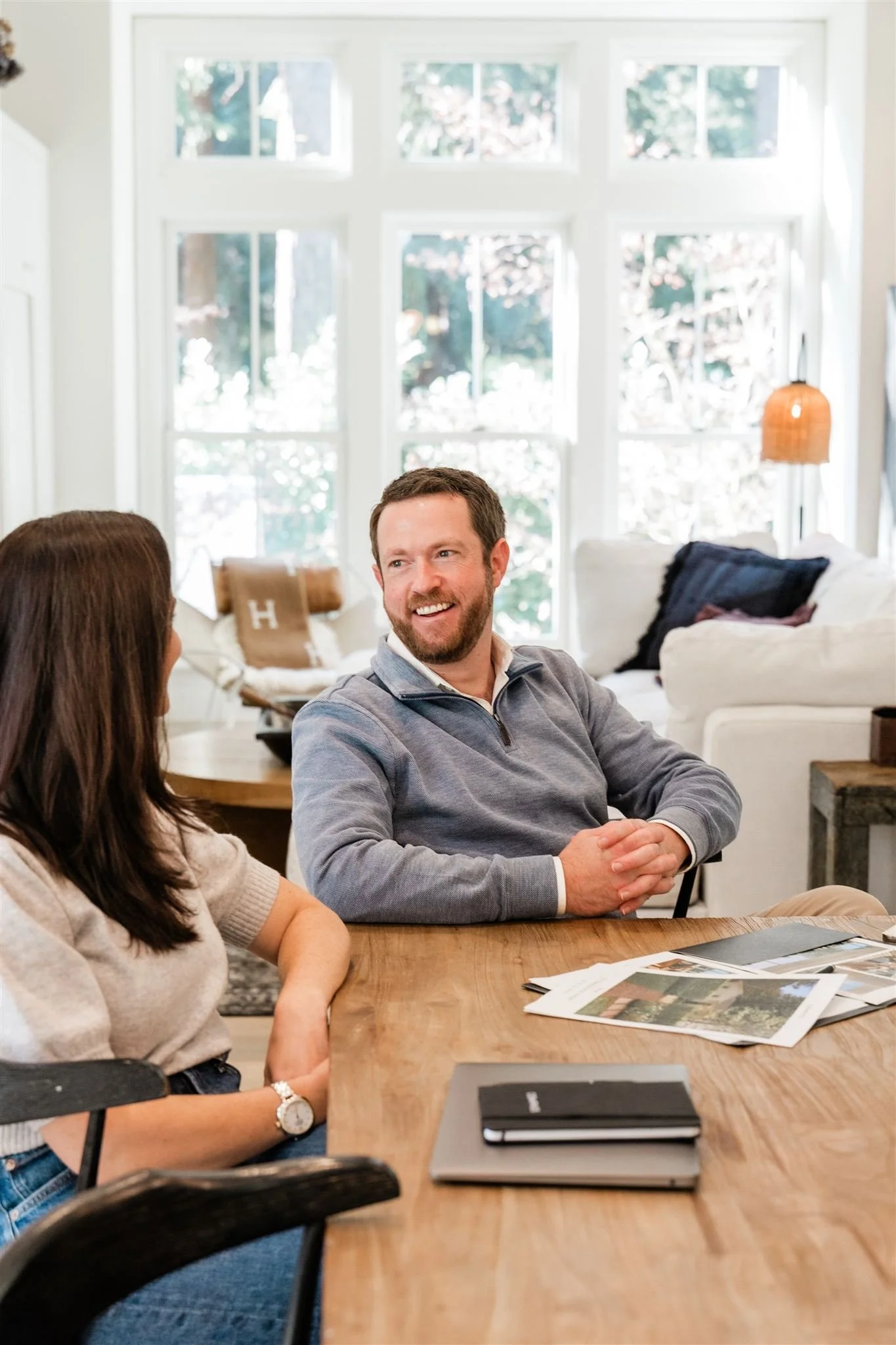 A man and woman sitting at a wooden table, engaged in conversation inside a bright room with large windows.