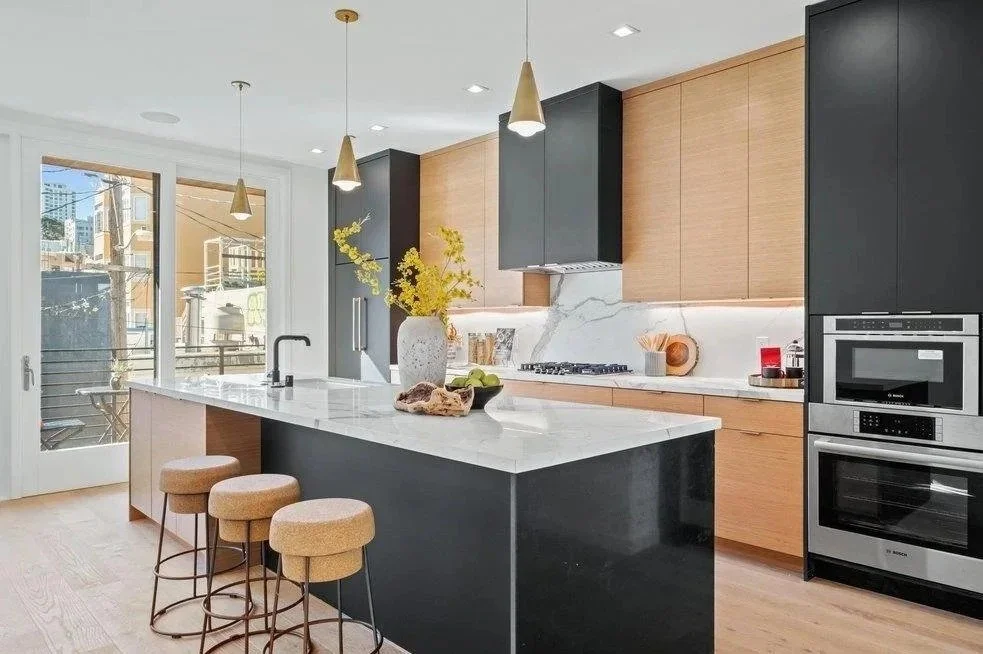 Modern kitchen with black and wooden cabinets, white marble countertop, and large windows with a city view. There are three beige bar stools at the island, and decorative items like a vase with yellow flowers and kitchen accessories on the counter.