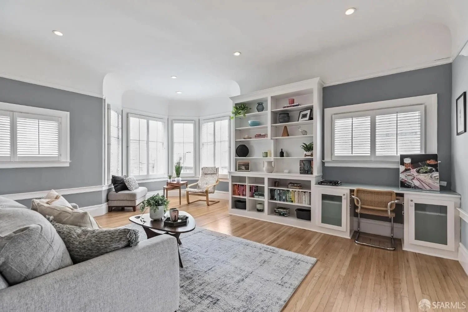 Bright living room with gray walls, large windows, white built-in shelves, a gray sofa with pillows, a coffee table with decor, an armchair, and a small desk with a TV.