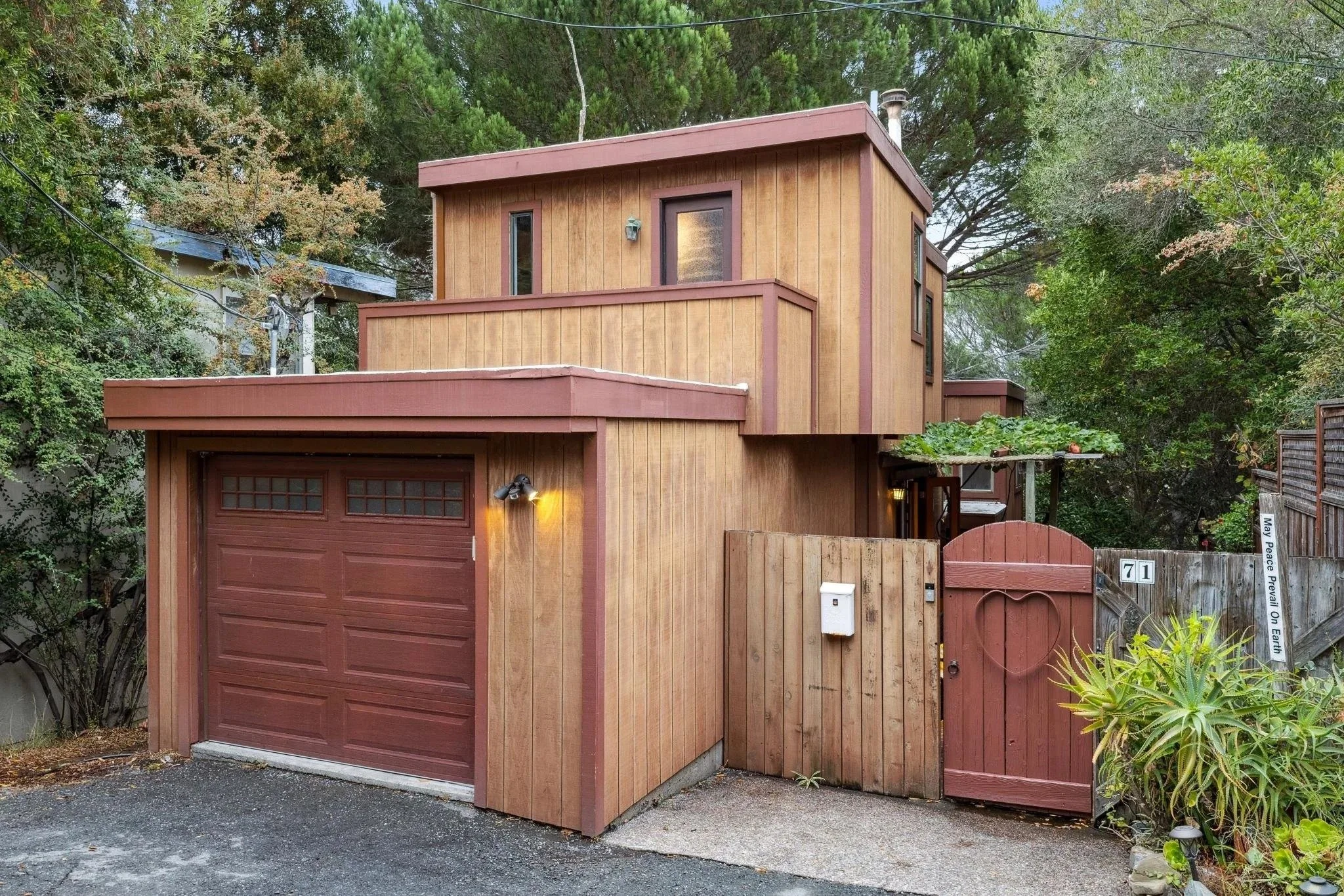 A modern, multi-story house with wooden siding, a garage door, and a gated entrance, surrounded by trees and greenery.