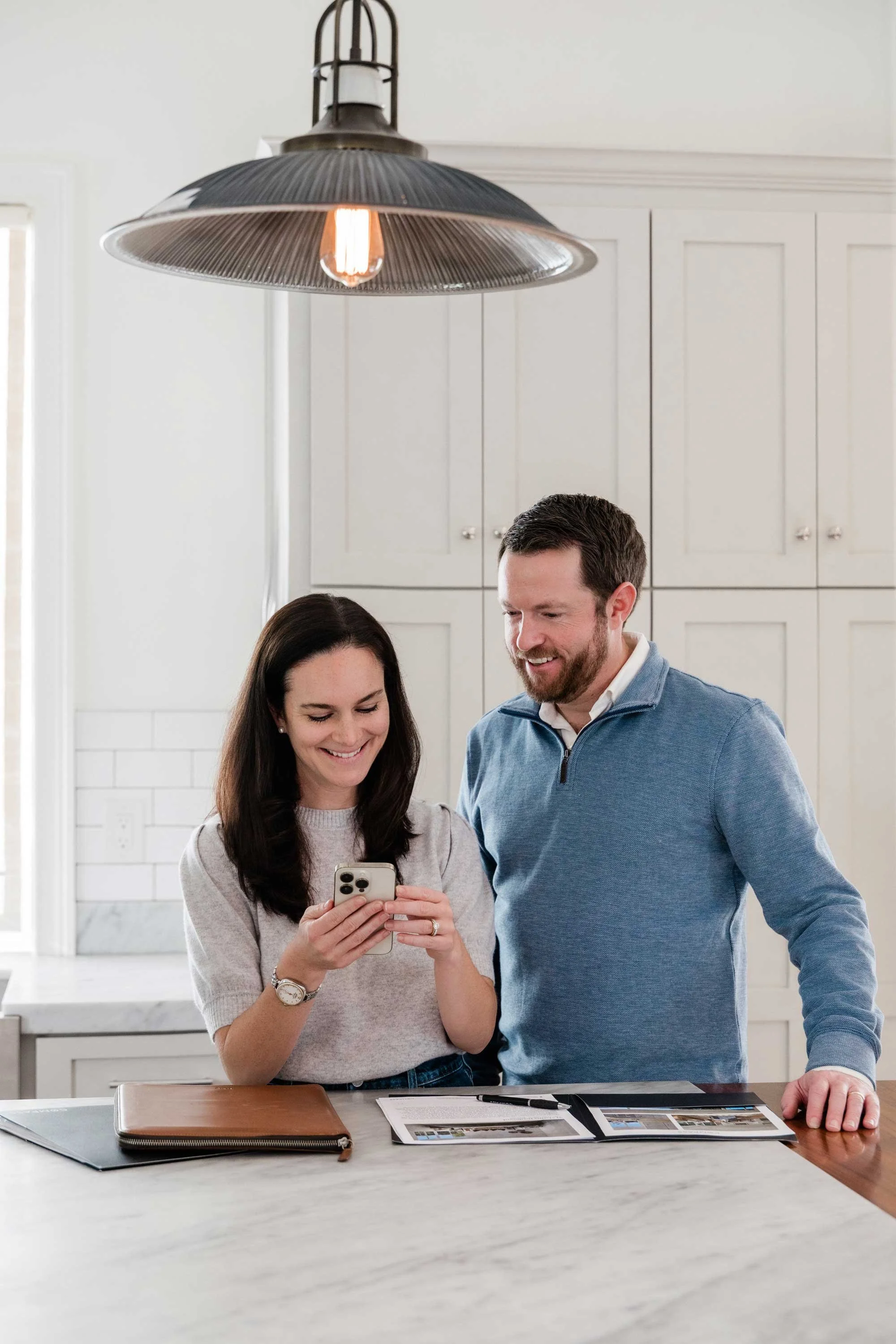 A woman and a man in a bright kitchen looking at a smartphone together, smiling.