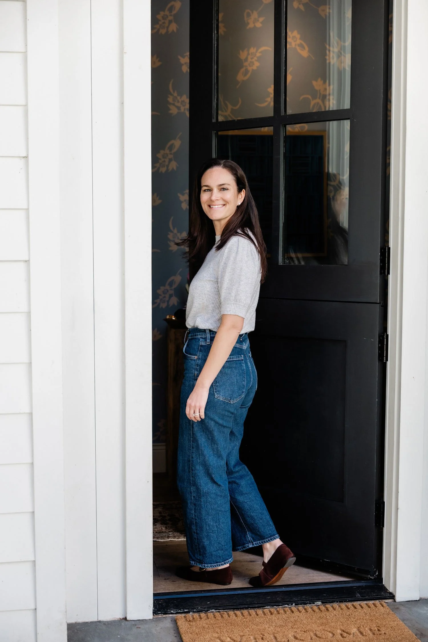 A woman with dark hair smiling, standing at an open black door in a house, wearing a grey top and blue jeans.