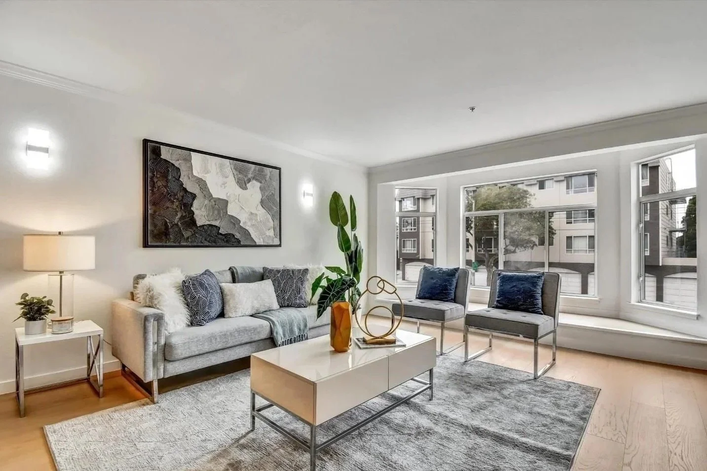 A modern living room with a light gray sofa, two gray armchairs with blue pillows, a white coffee table with a large green plant and decorative sculpture, a side table with a lamp and plant, a large abstract black and white painting, and a large bay window with views of a building and trees.