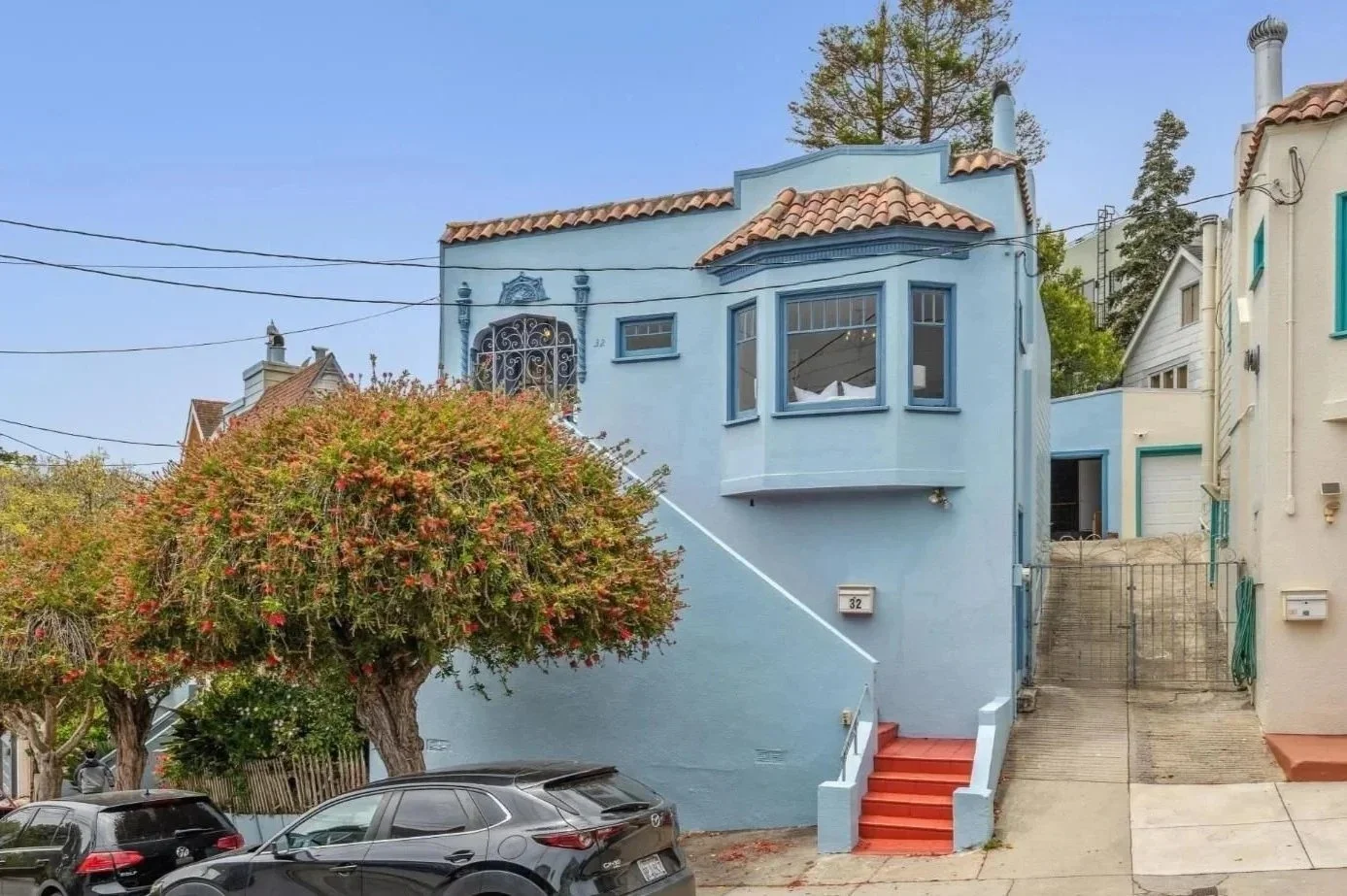 A blue house on a hill with stairs and a tree in front, parked cars below, and a clear sky.