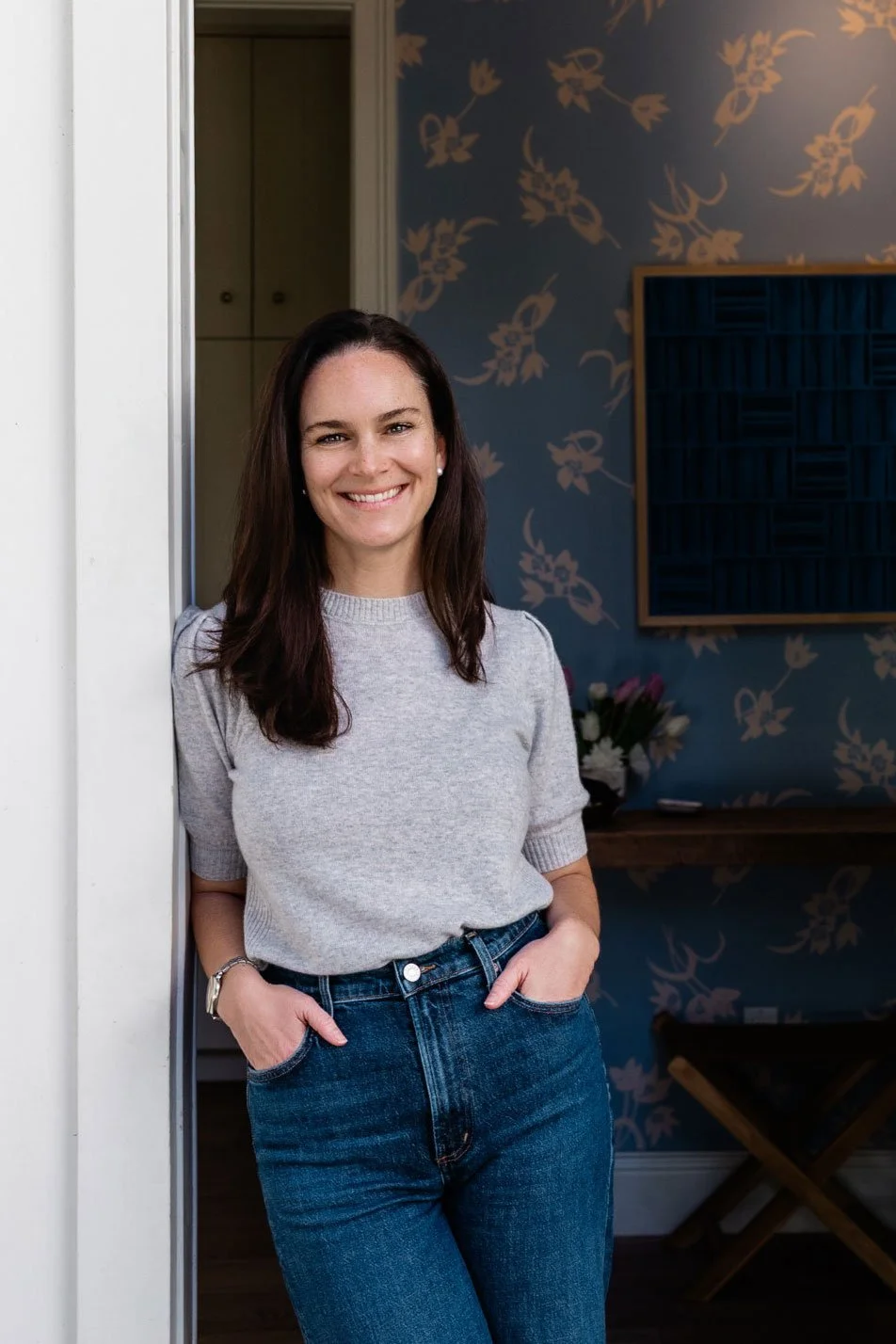 A woman with long dark hair and a light gray sweatshirt standing in an interior space with floral wallpaper, smiling, with her hands in her jeans pockets.