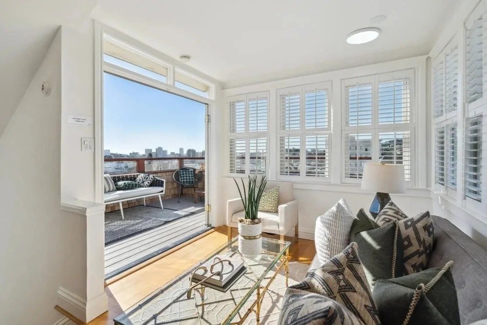 Living room with large windows, white shutters, and balcony with outdoor seating, including a bench and two chairs, overlooking a cityscape.