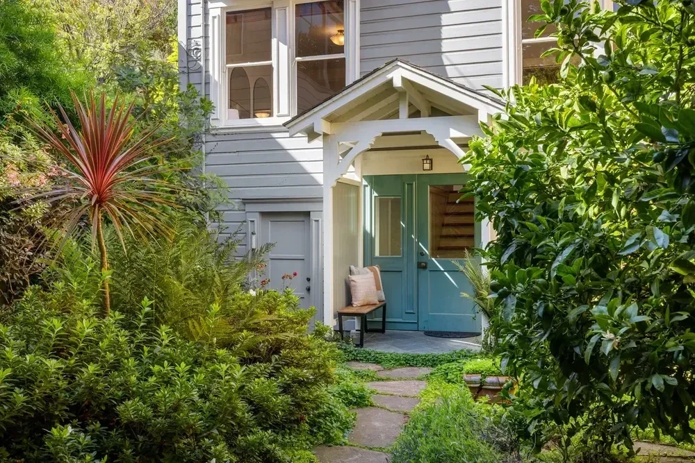 Blue front door with white trim and a small porch, surrounded by lush green plants and bushes, with a stone pathway leading to the entrance.