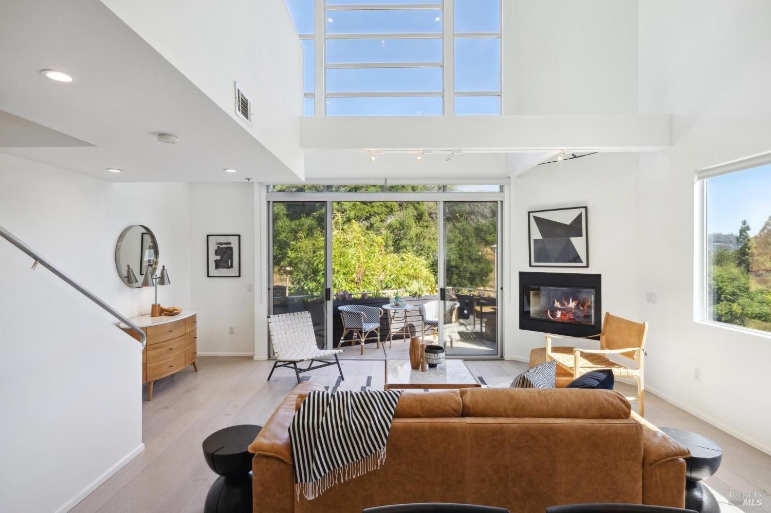Bright living room with large windows and sliding glass door opening to a patio with outdoor furniture, modern artwork, fireplace, and tan leather sofa, with white walls and light wood flooring.