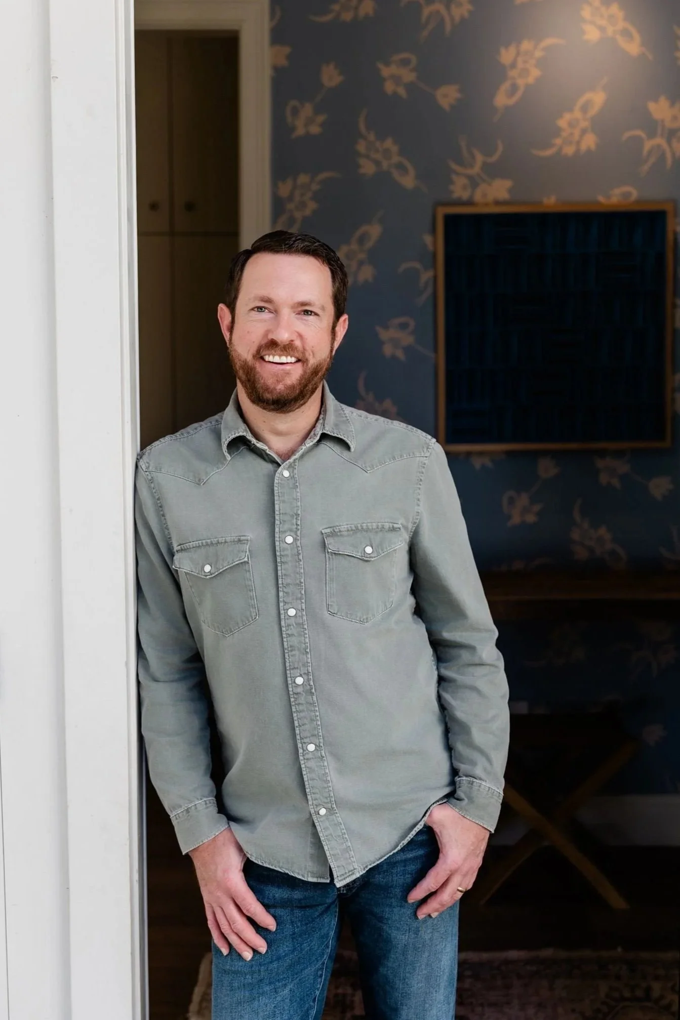 A man with a beard, wearing a light gray button-up shirt and blue jeans, smiling and standing in a doorway.