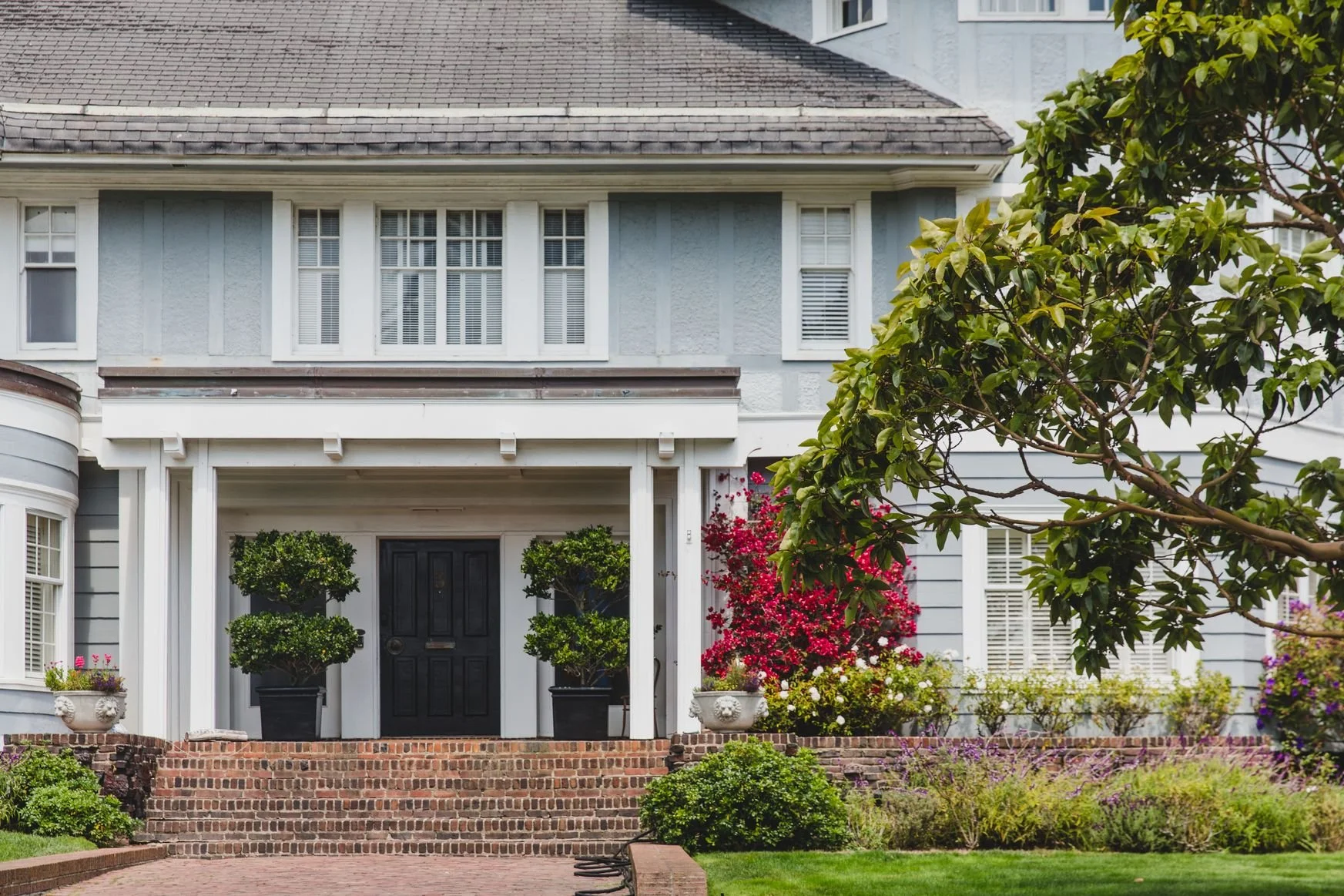 Front view of a light blue house with white trim, a black front door, potted topiary plants, and colorful flowering bushes, including pink and white blossoms. A tree partially obscures the right side of the house.