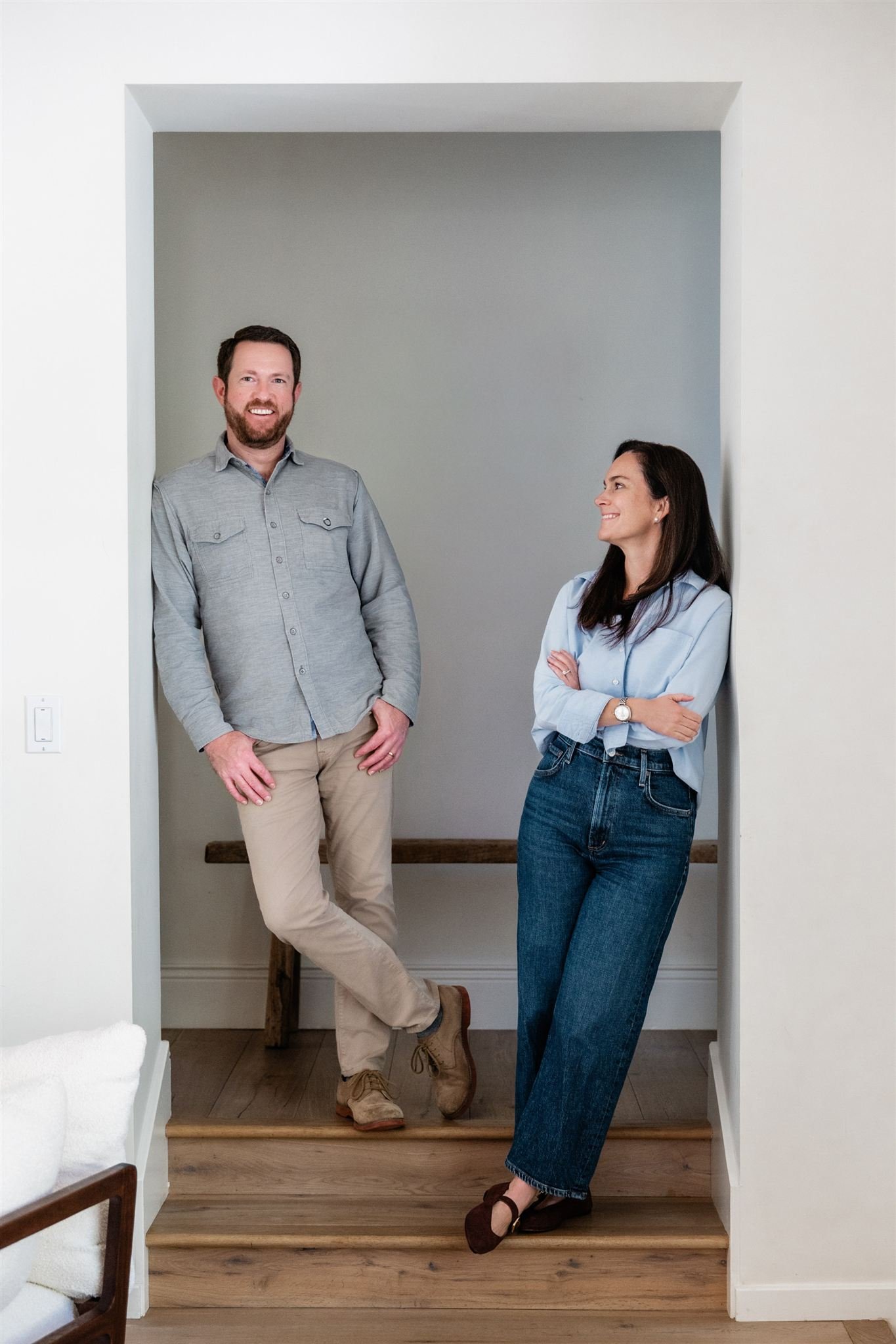 A man and woman standing in a small nook, smiling and looking at each other, with a wooden bench behind them.