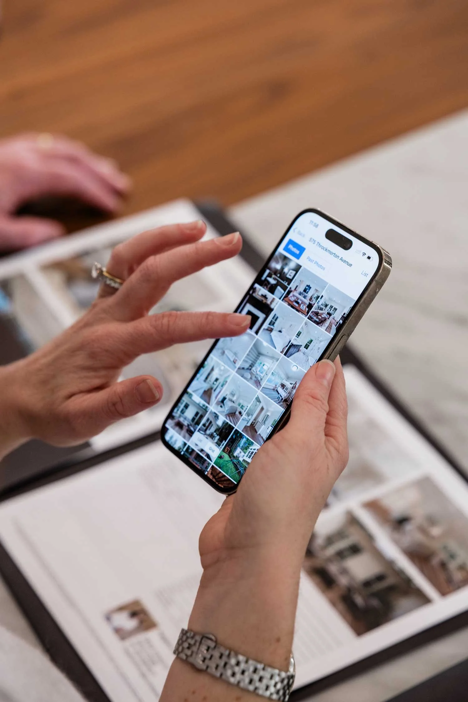 Person using a smartphone to browse a real estate app showing images of home interiors on a table with open magazines.