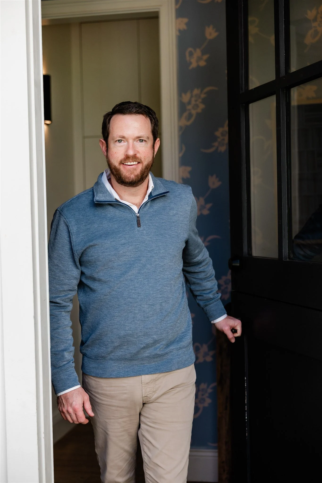 A man with a beard and short dark hair, wearing a blue quarter-zip sweater and beige pants, standing in a doorway.