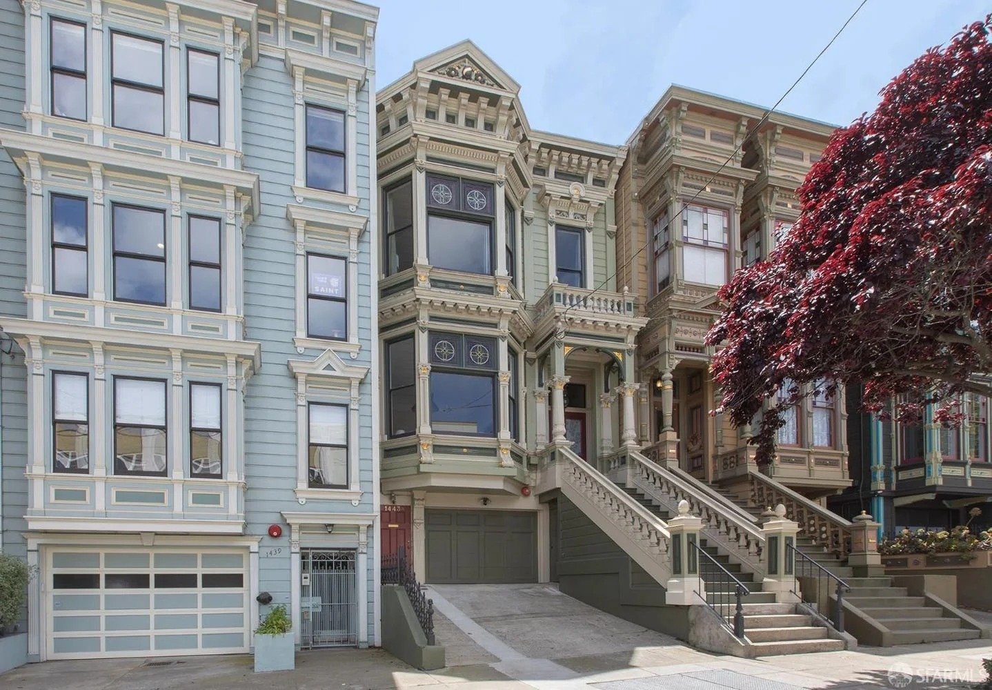 Three Victorian-style row houses in San Francisco with colorful facades, stairs, and bay windows, beside a large red-leafed tree.