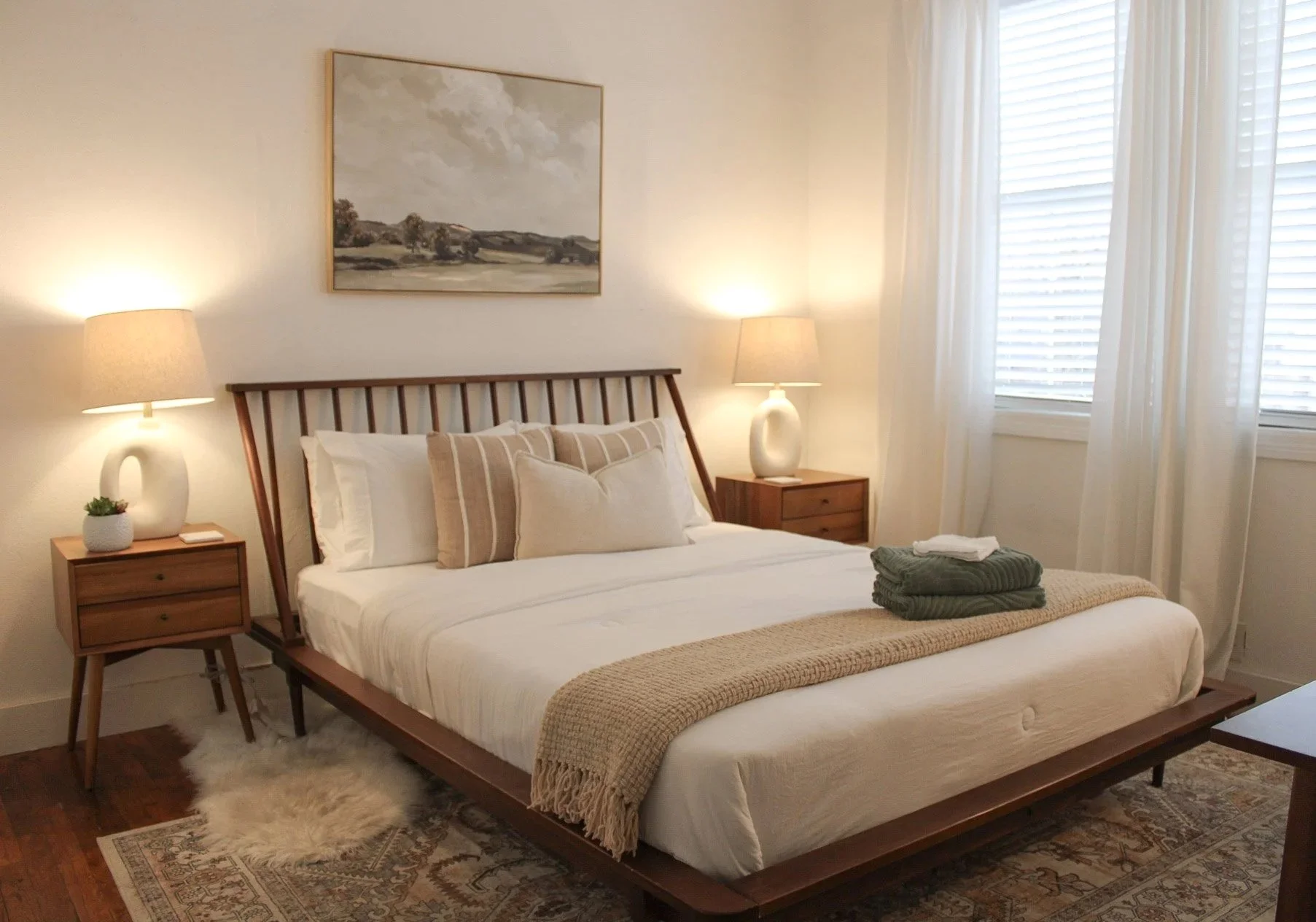 A neatly made bed with white and beige pillows, a beige throw blanket, and folded towels on top. There are two matching wooden nightstands with lamps, a small potted plant, and a painting of a landscape hanging above the bed. The room has white walls