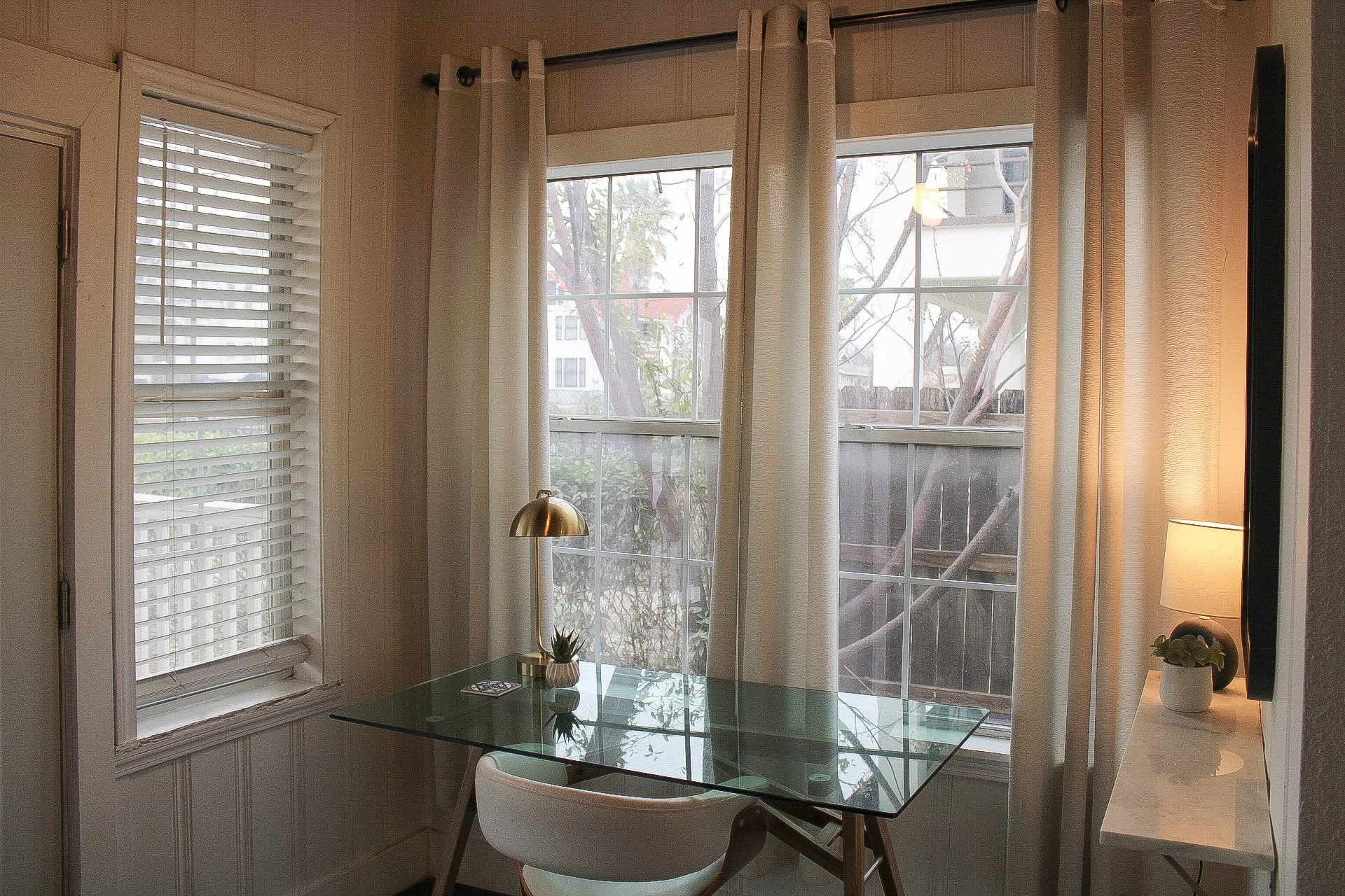 A small home office with a glass desk, a white chair, and a window with white curtains. The room has wood-paneled walls and a side table with a lamp and a potted plant.