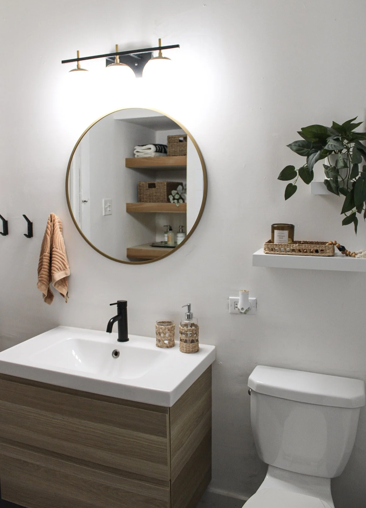 A modern bathroom with a white sink, wooden vanity, black faucet, round mirror, white toilet, wall-mounted shelf with plant and decor, towel hooks with a peach towel, and shelf with baskets and decorative items.