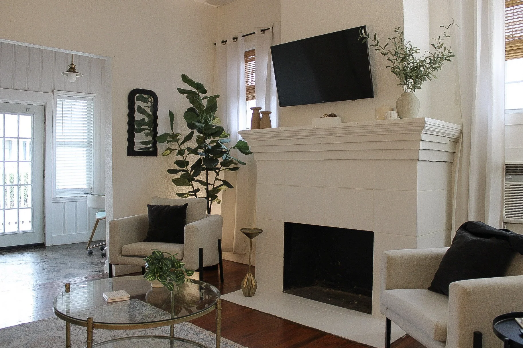 Living room with white walls, a white fireplace, a wall-mounted TV above the fireplace, potted plants, armchairs with black pillows, a glass coffee table, and a door and windows with white curtains letting in natural light.
