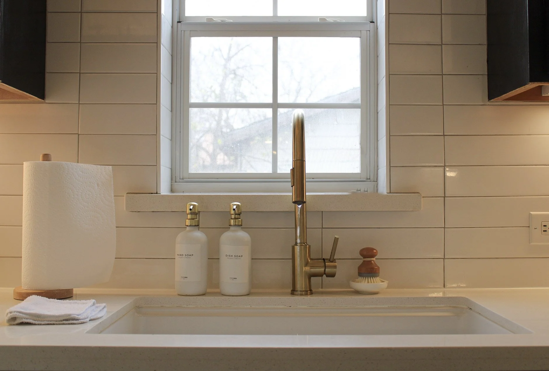 Kitchen sink area with a window, gold faucet, soap bottles, paper towel roll, dish brush, and folded towel on the countertop.