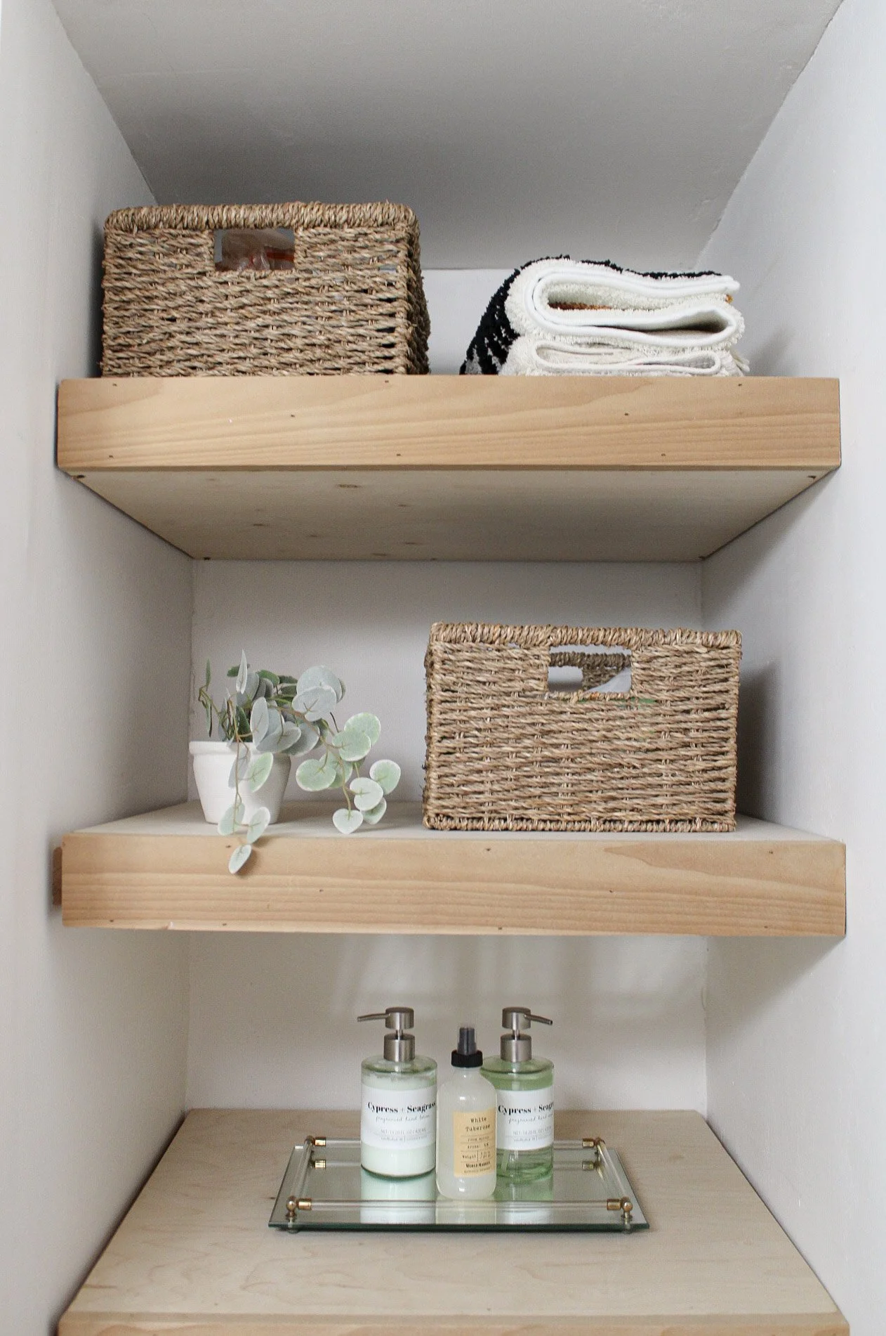 Decorative shelves with woven baskets, folded towels, a small potted plant, and bottles of liquid soap or lotion on a mirrored tray.