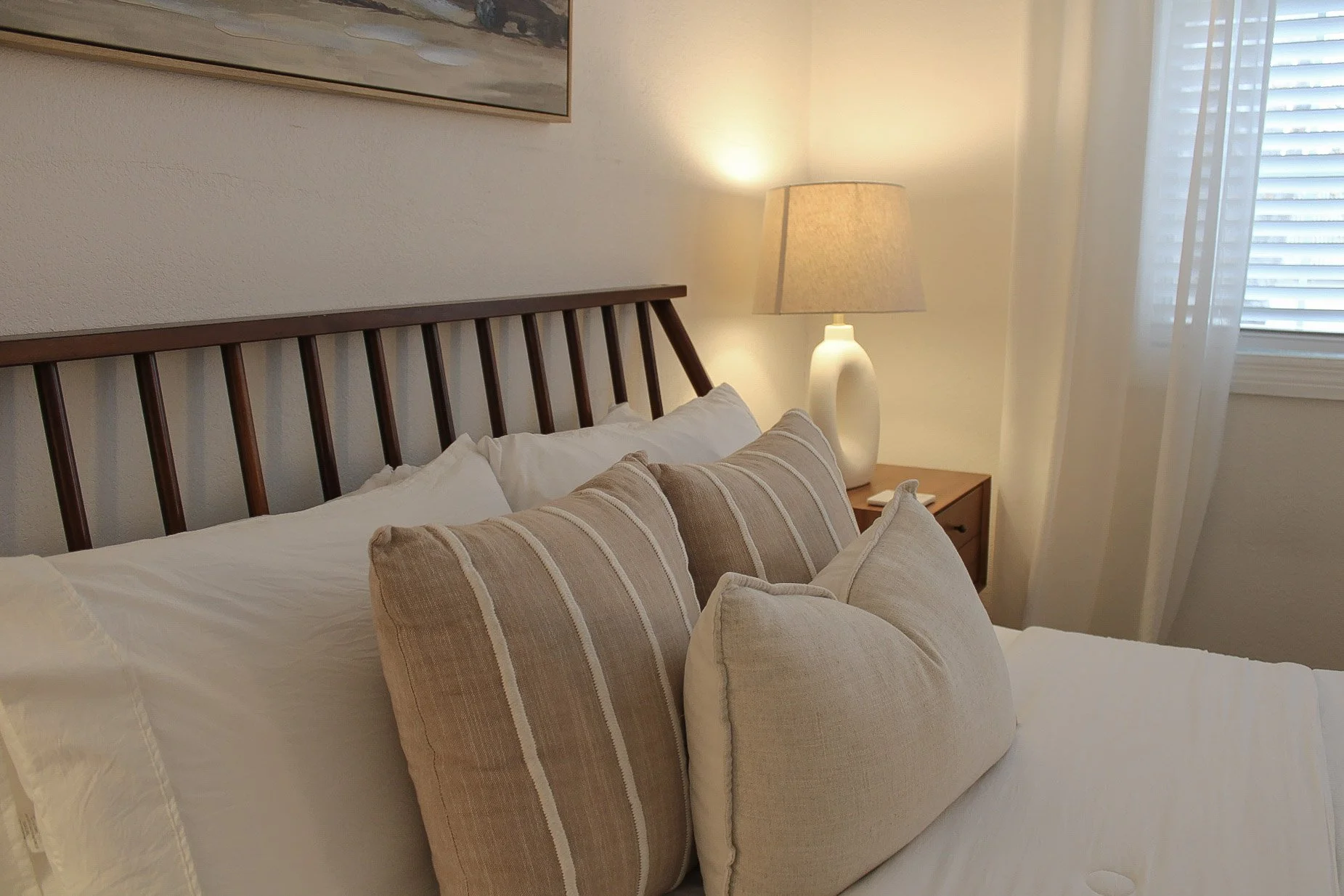 A neatly made bed with white sheets, multiple pillows including a large striped pillow, a wooden headboard, a bedside table with a white ceramic lamp, and a window with blinds and curtains.