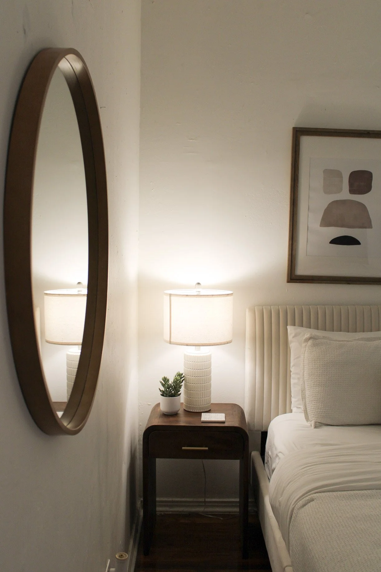 A bedroom corner with a wooden nightstand, two table lamps, a small potted plant, and a framed abstract art piece on the wall.