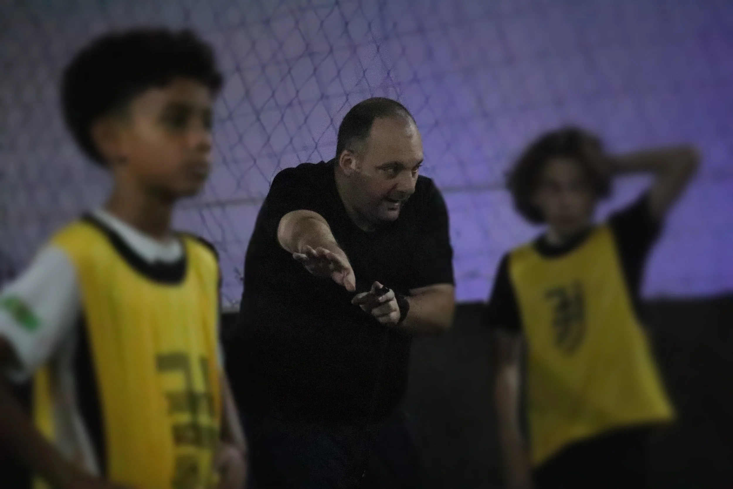 Professor ou treinador orientando duas crianças durante uma atividade esportiva indoor, com uma rede ao fundo.