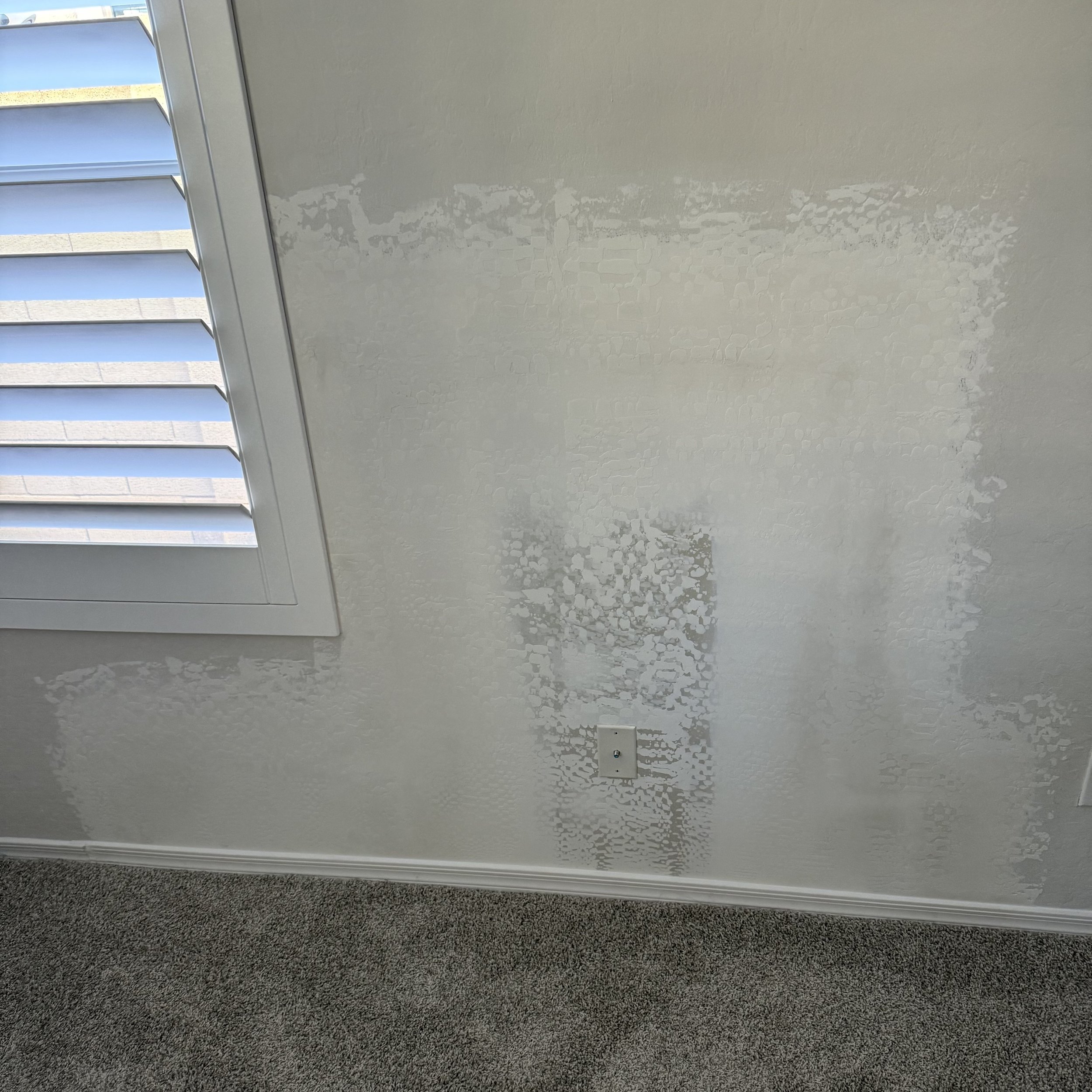 A wall with patches of textured white paint or plaster around an electrical outlet, with a window with white shutters on the left side, and a carpeted floor at the bottom.