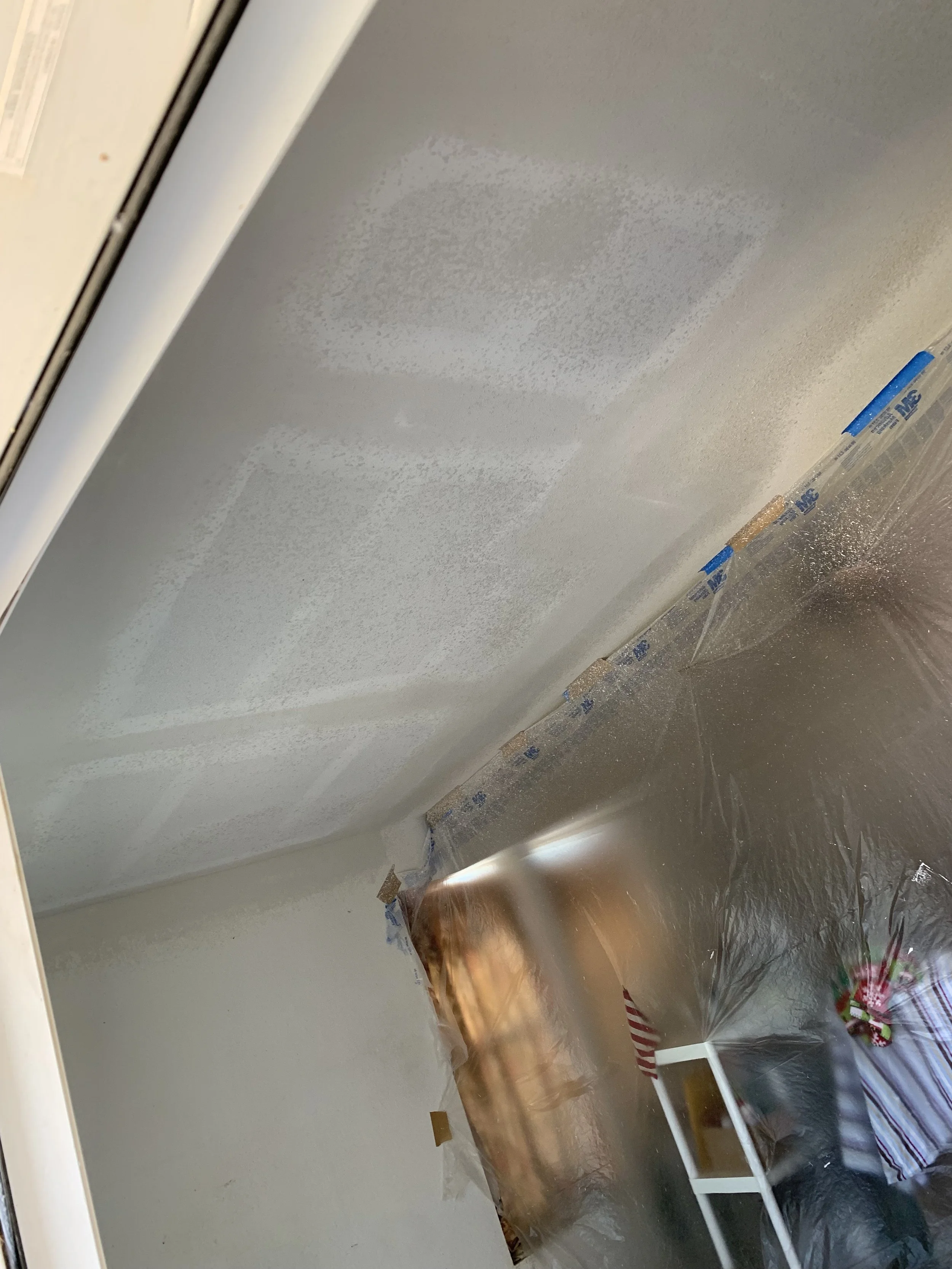 A ceiling with three patches of primer or paint, visible from the dried spots. The image captures part of a room with a plastic sheet covering furniture and a nearby window, likely during a painting or renovation process.