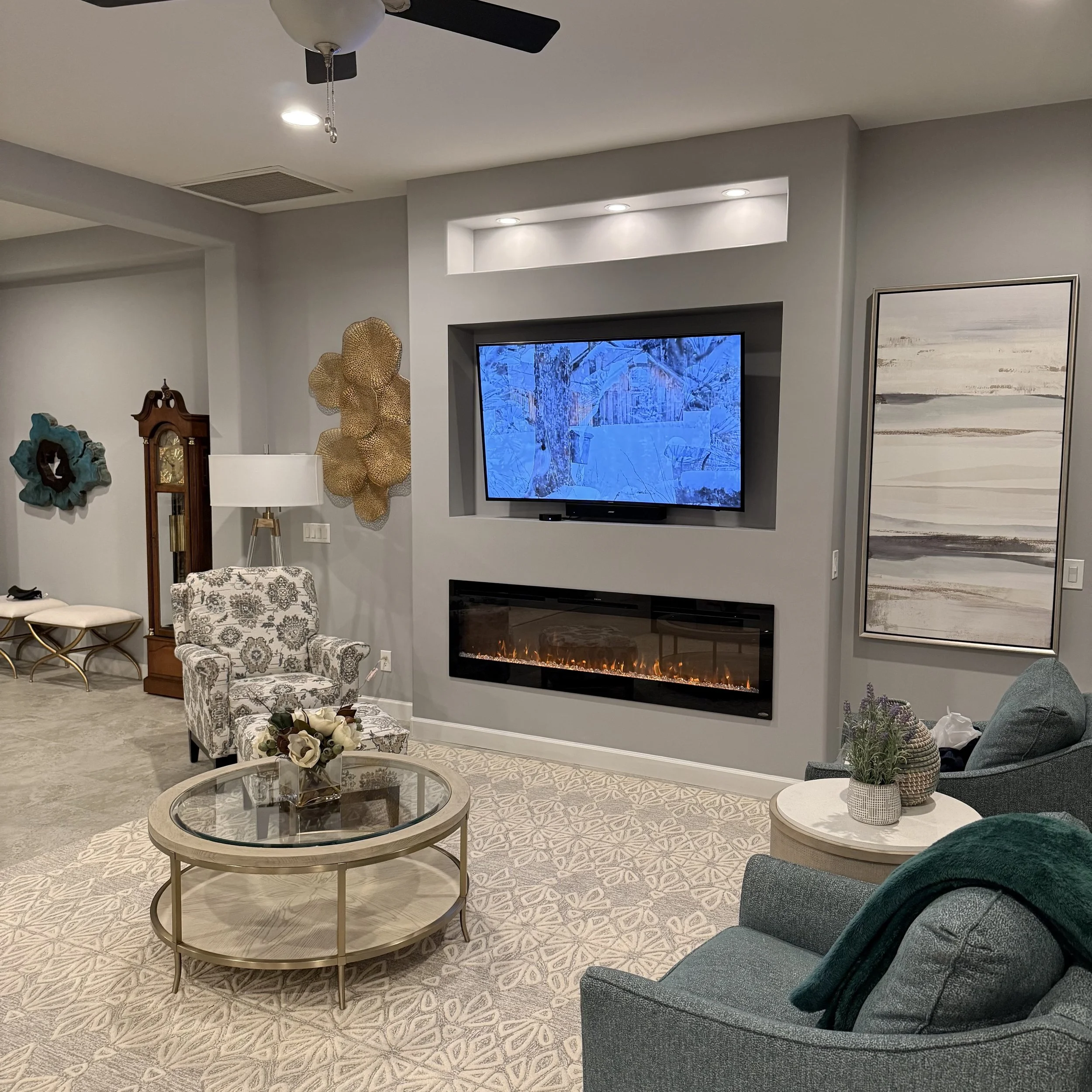 Living room with a gray wall featuring a mounted flat-screen TV displaying a snowy scene, a modern electric fireplace below, a patterned carpet, a glass coffee table with a floral arrangement, a comfortable armchair with a blanket, a side table holding decorative vases, an upholstered chair, a tall grandfather clock, decorative wall pieces, and a ceiling fan with lights.