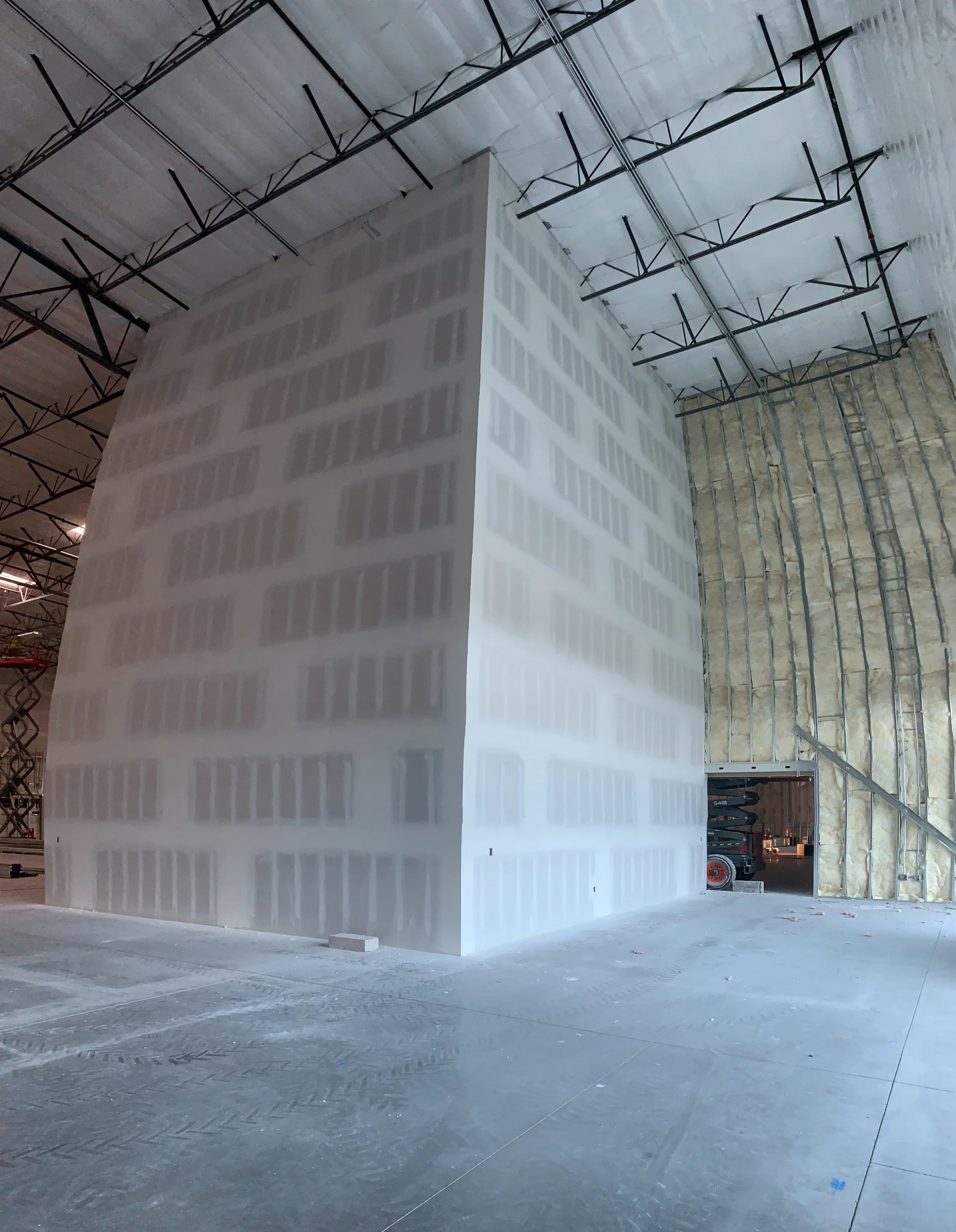 A large construction site featuring a tall, partially finished drywall structure with visible joints, inside an industrial building with exposed ceiling beams and insulation on one wall.
