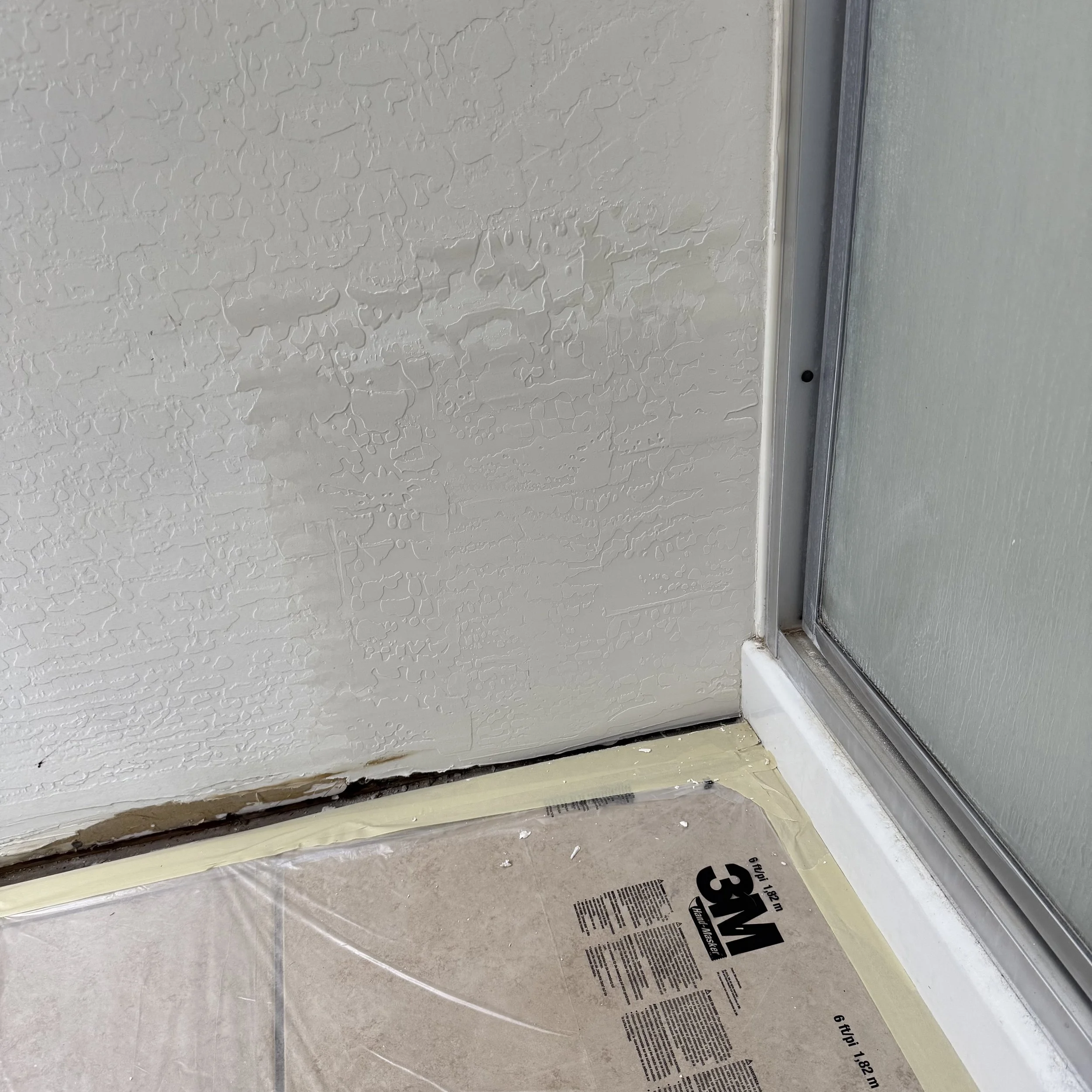 Close-up of a textured white wall and a glass shower door with a metal frame. The bottom of the wall has painter's tape and a piece of wallpaper protection paper on the floor.