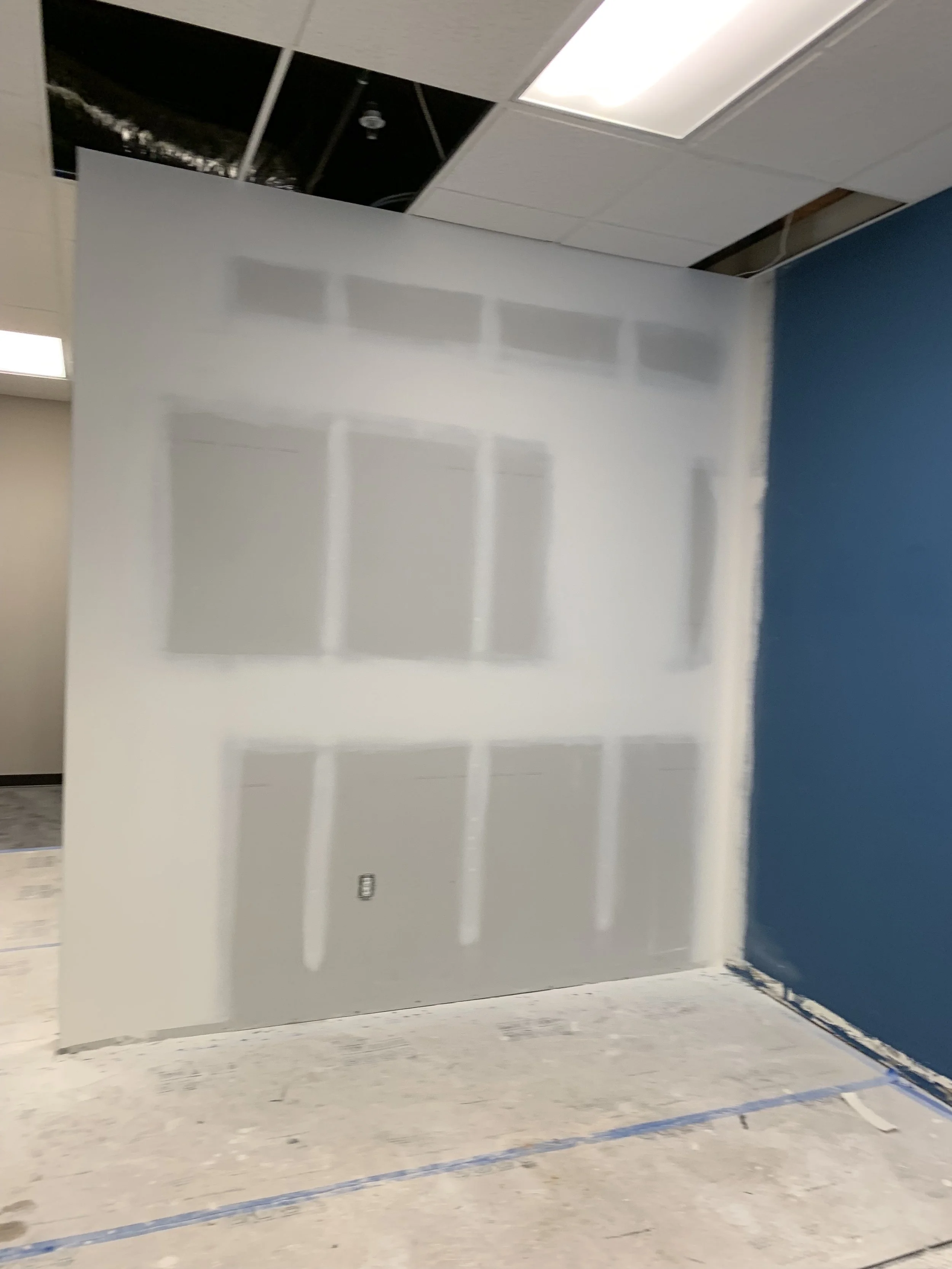 Partially completed drywall installation in an office or commercial space, with drywall panels and painting in progress, exposed ceiling panels, and protective tape on the floor.