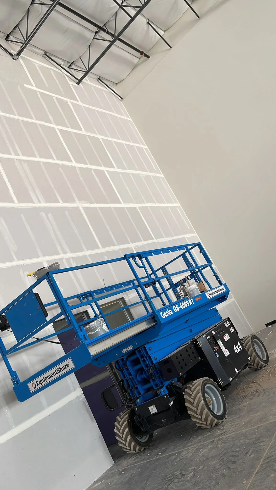 Blue Genie GS-4069 RT aerial work platform inside a large, unfinished room with white walls and marked flooring. The platform has large tires and raised railing.