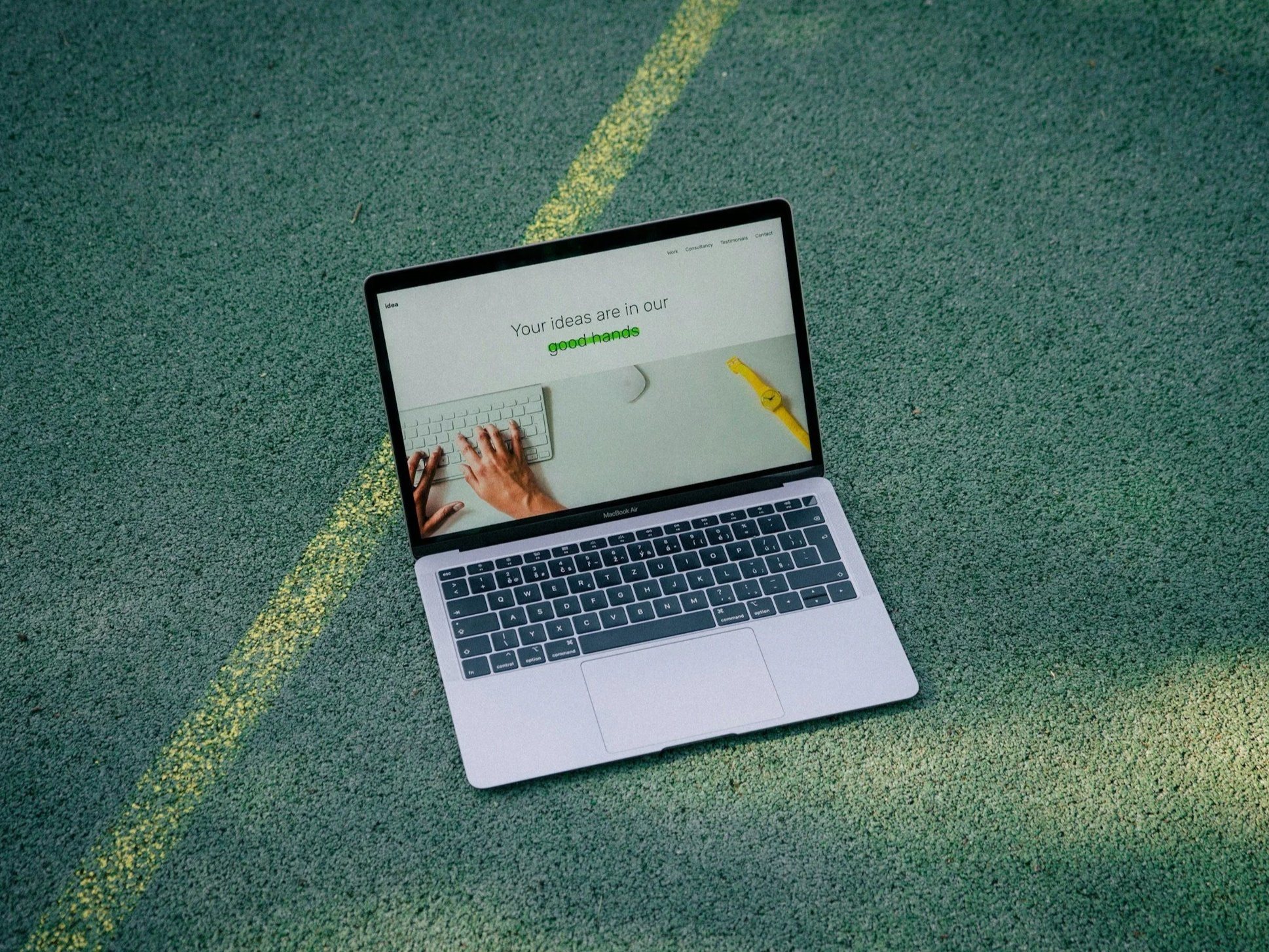 A laptop on a wooden table displaying a website about interior design, with a man in a white shirt smiling on the screen, and some indoor plants partially visible in the foreground.