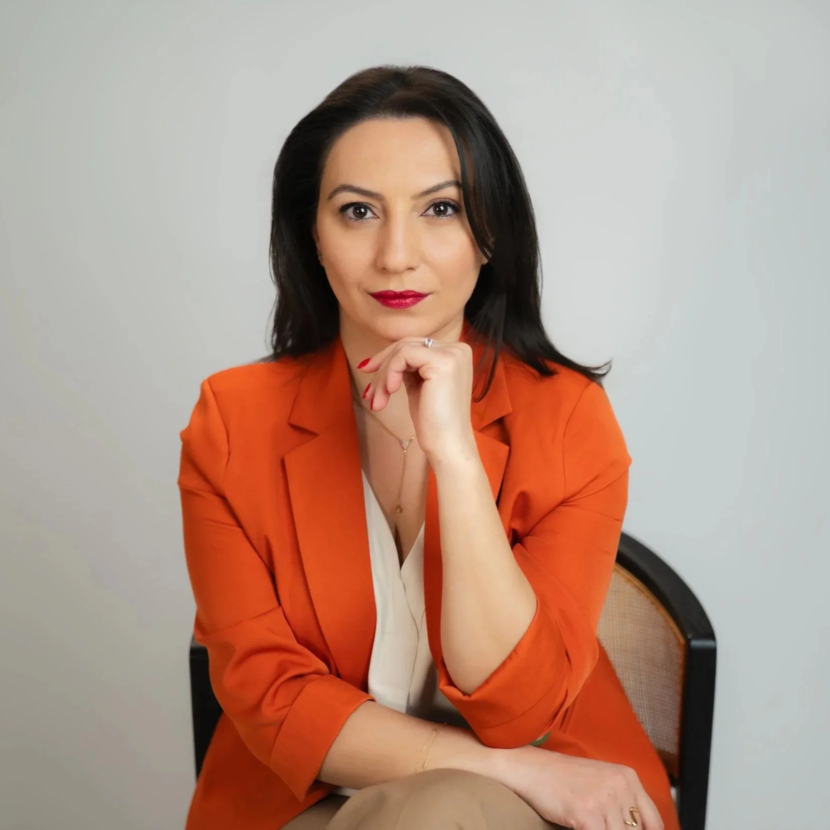 A woman with black hair wearing a red lipstick and a professional orange blazer sitting in an office chair in front of a plain white wall. The woman is Stella Grigoryan, the founder of ANALOGIA Creative Agency.