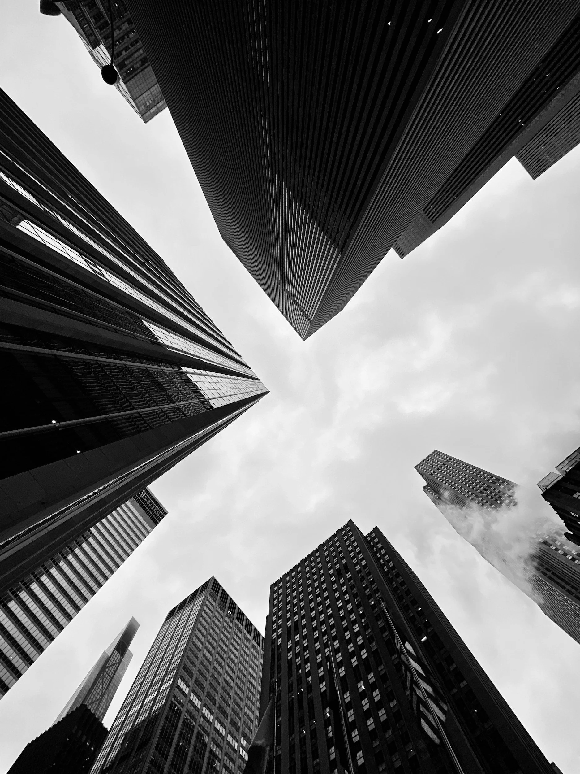 Black and white photo of towering skyscrapers viewed from the ground looking up at the cloudy sky.