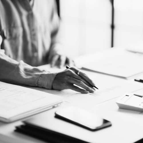 Close-up of a person's hand writing or reviewing documents on a desk with a smartphone, laptop, and office supplies in a bright office setting.