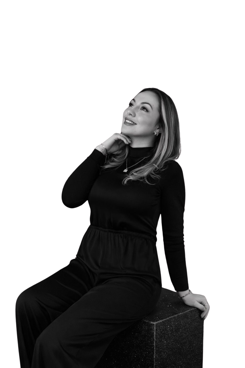 A woman with light-colored hair, wearing a black long-sleeve top and black pants, sitting on a black textured cube, looking up with her hand resting on her chin.