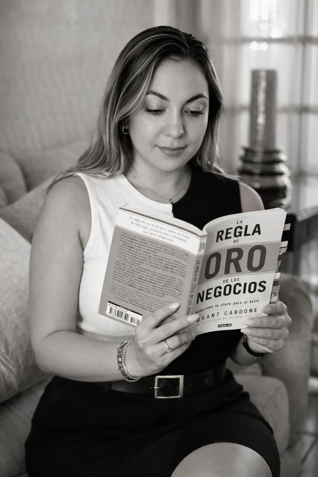 A woman with long hair reads a book titled "La Regla de Oro de los Negocios" in a living room.