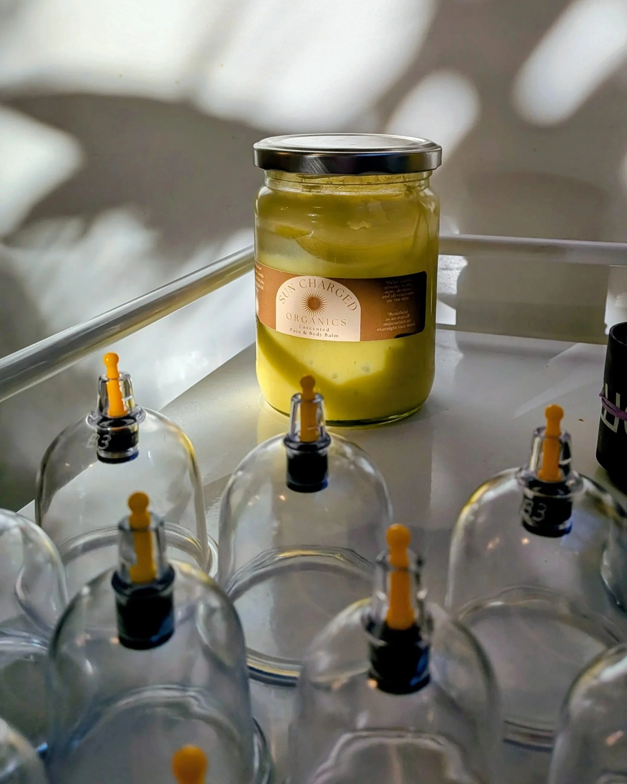 A jar of Sun Charged Organics undereye and face balm on a white surface with myofascial cupping cups in the foreground. Used for massage, trigger point therapy, soft tissue, cupping, relaxation massage and lymphatic massage in aid of injuries