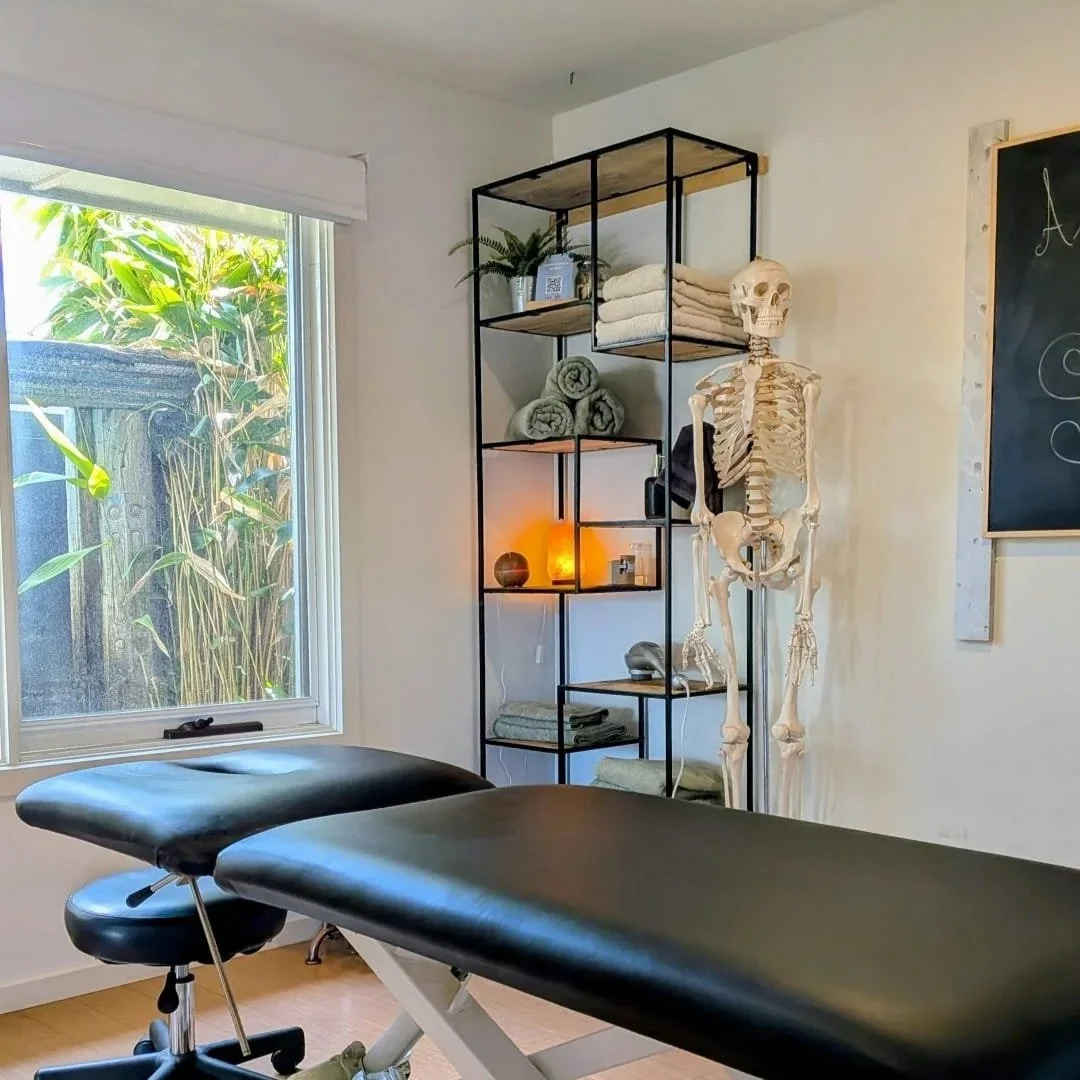 Interior of a massage or therapy room with a massage table, a black rolling stool, a window with green plants outside, a metal shelving unit with towels and decor, and a human skeleton model beside the shelf located in Ocean Grove on the Bellarine