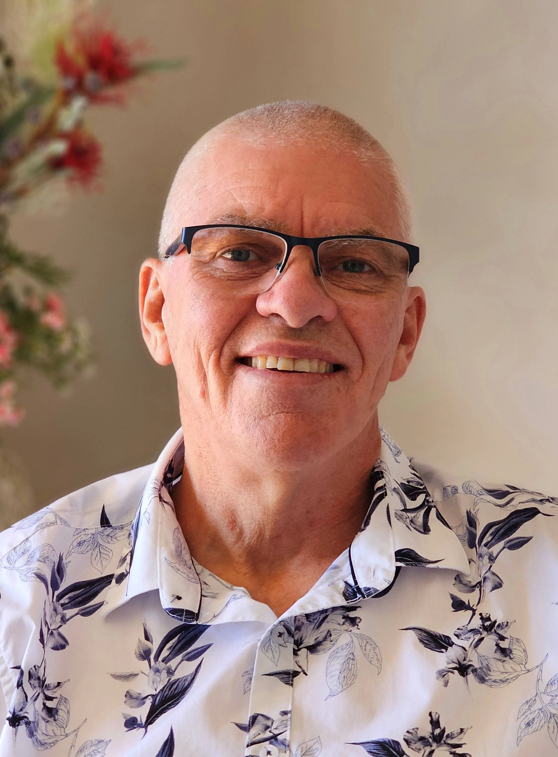 A smiling man with short gray hair, glasses, wearing a white shirt with black floral pattern, standing indoors with blurry plant in background.