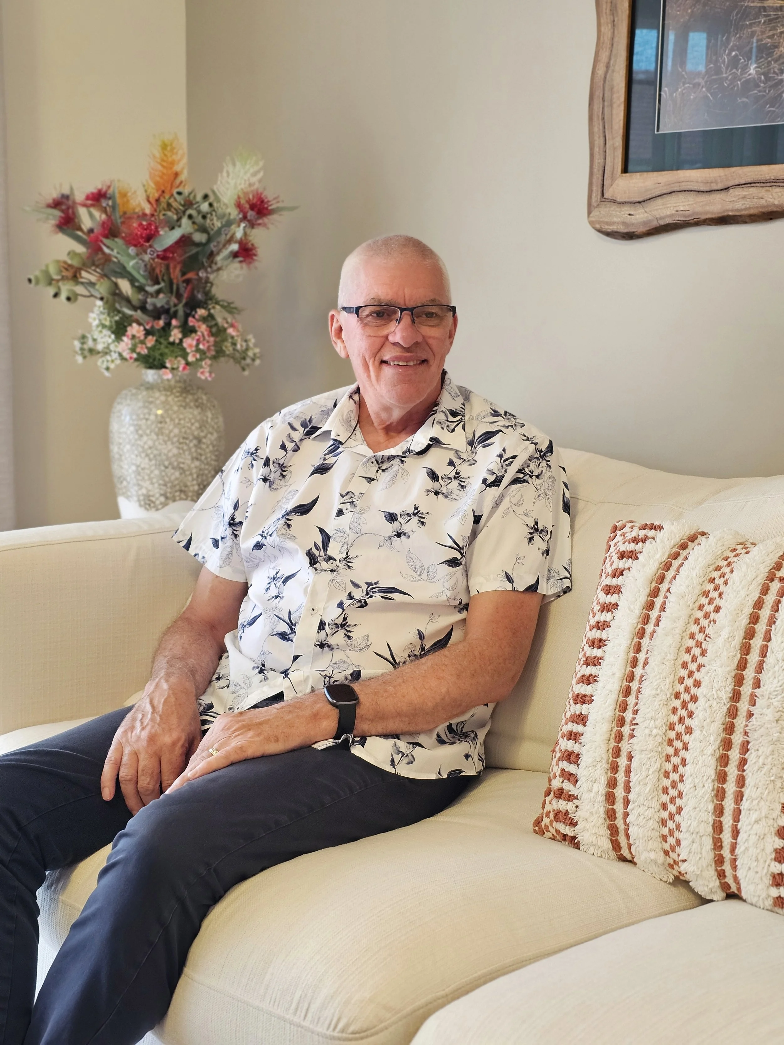 A man with glasses, wearing a floral short-sleeve shirt and a smartwatch, sitting on a cream-colored sofa in a living room with a floral arrangement on a side table and a framed picture on the wall.