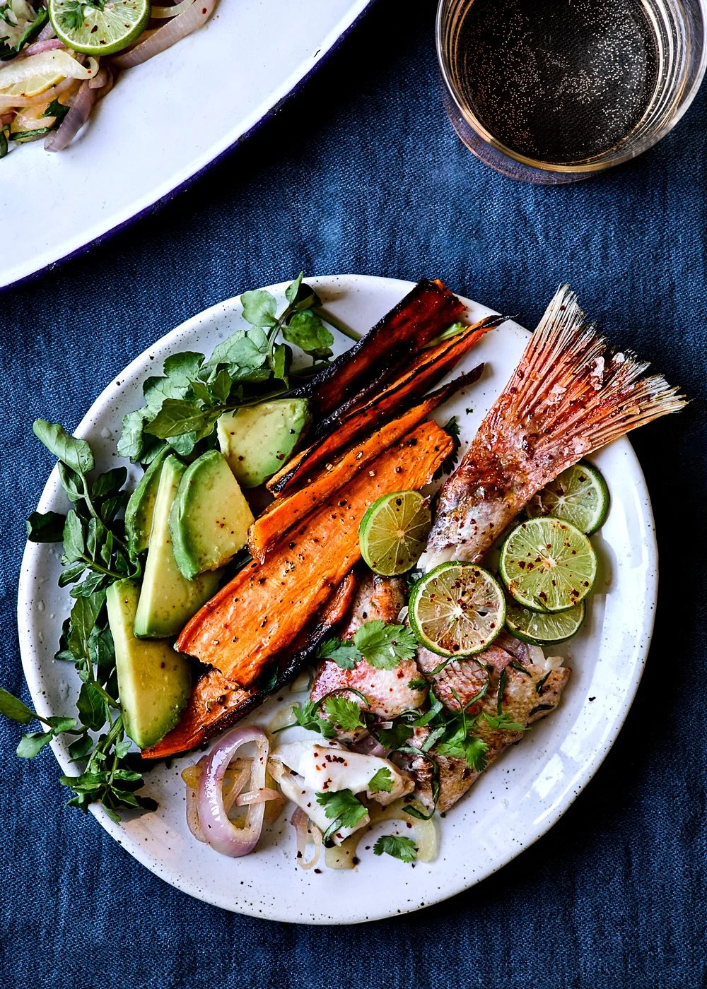A plate of grilled fish with sliced limes, avocado, grilled vegetables, and greens on a blue tablecloth, with a glass of dark soda nearby.