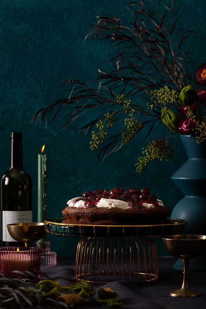 A chocolate cherry cake with whipped cream and cherries on top, on a gold cake stand, surrounded by candles, a bottle, and a large vase with flowers against a dark green wall.